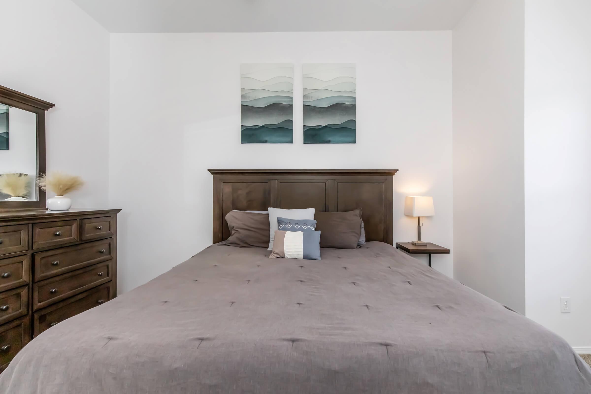 A cozy bedroom featuring a dark wooden bed frame with a neutral-colored comforter. A small lamp is placed on a nightstand next to the bed. A dresser with multiple drawers is on one side, and two abstract landscape paintings in shades of blue and green are hung above the bed.