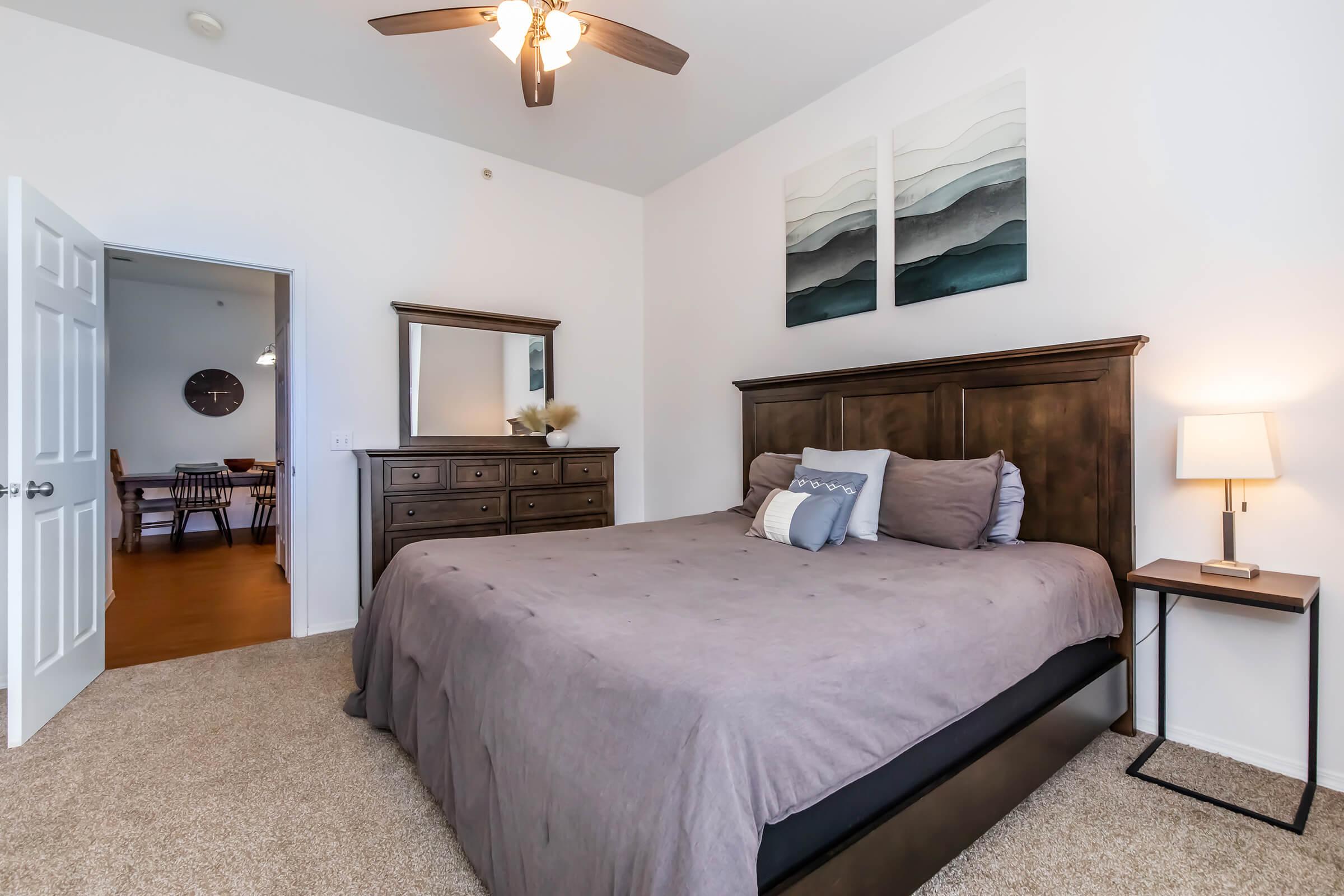 A cozy bedroom featuring a dark wooden bed frame with a gray comforter, matching nightstand, and dresser. The walls are painted white, and two abstract wall art pieces in shades of blue and gray are above the bed. A doorway leads to a dining area in the background, with warm wooden floors.