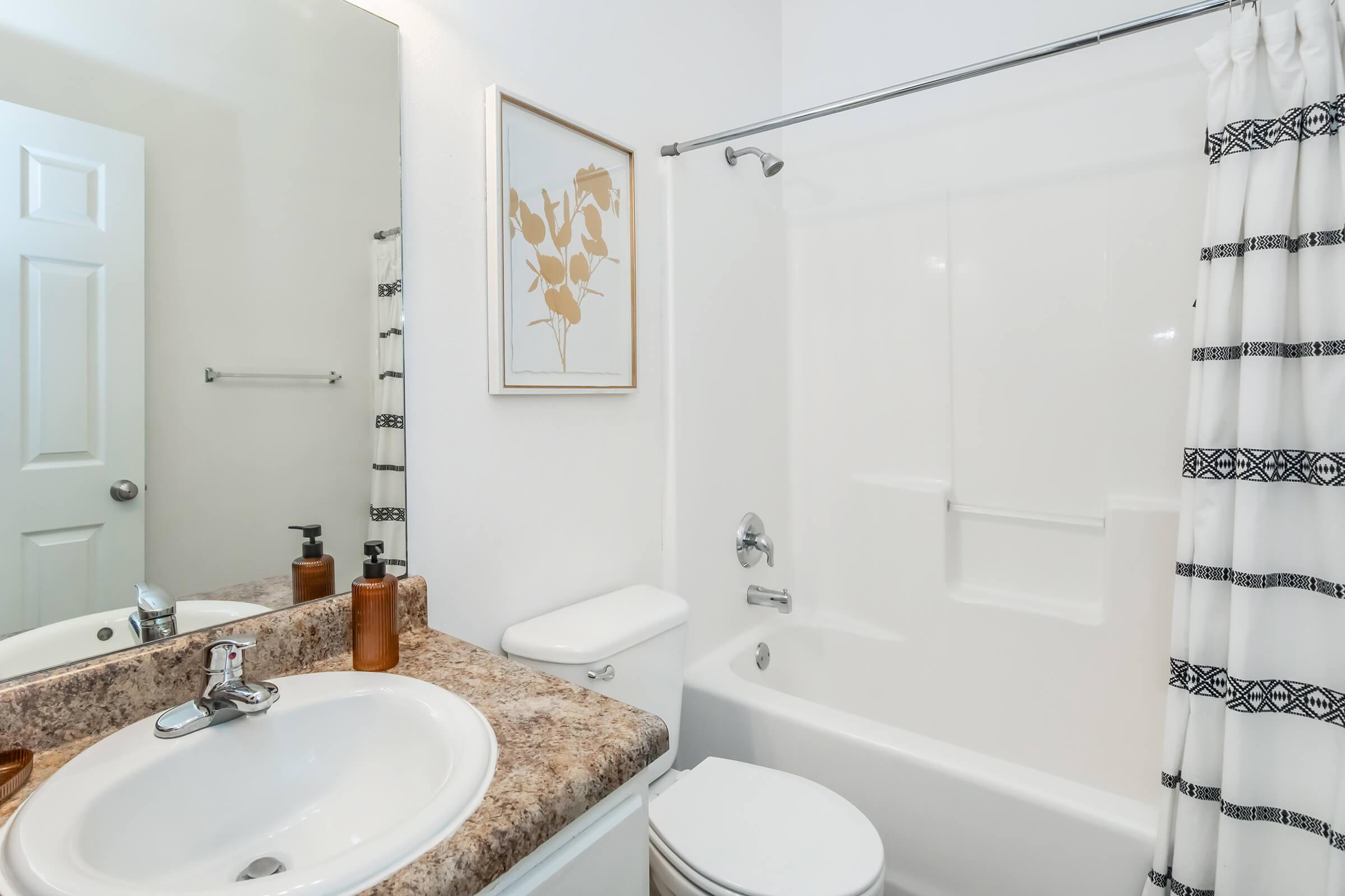 A clean and modern bathroom featuring a white bathtub with a shower curtain, a sink with a faucet, a wall-mounted mirror, and a decorative framed wall art. The countertop is made of speckled brown stone, and there are minimal toiletries visible on the sink. The walls are painted white.