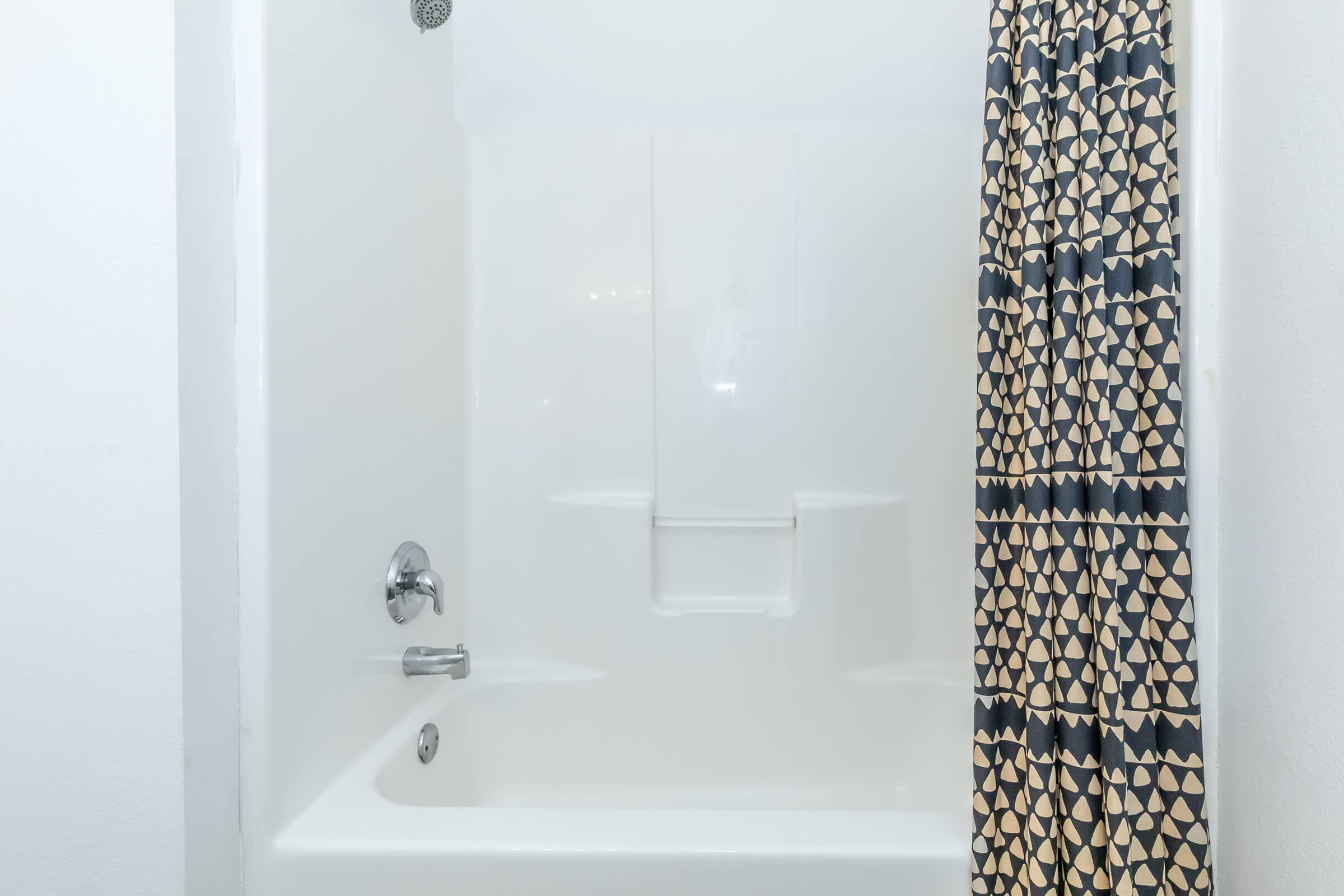 A clean, modern bathtub with a white finish. The bathtub features a chrome faucet and a showerhead. A patterned shower curtain with geometric designs is hanging from a rod, adding a decorative touch to the bathroom space. The overall ambiance is bright and minimalist.
