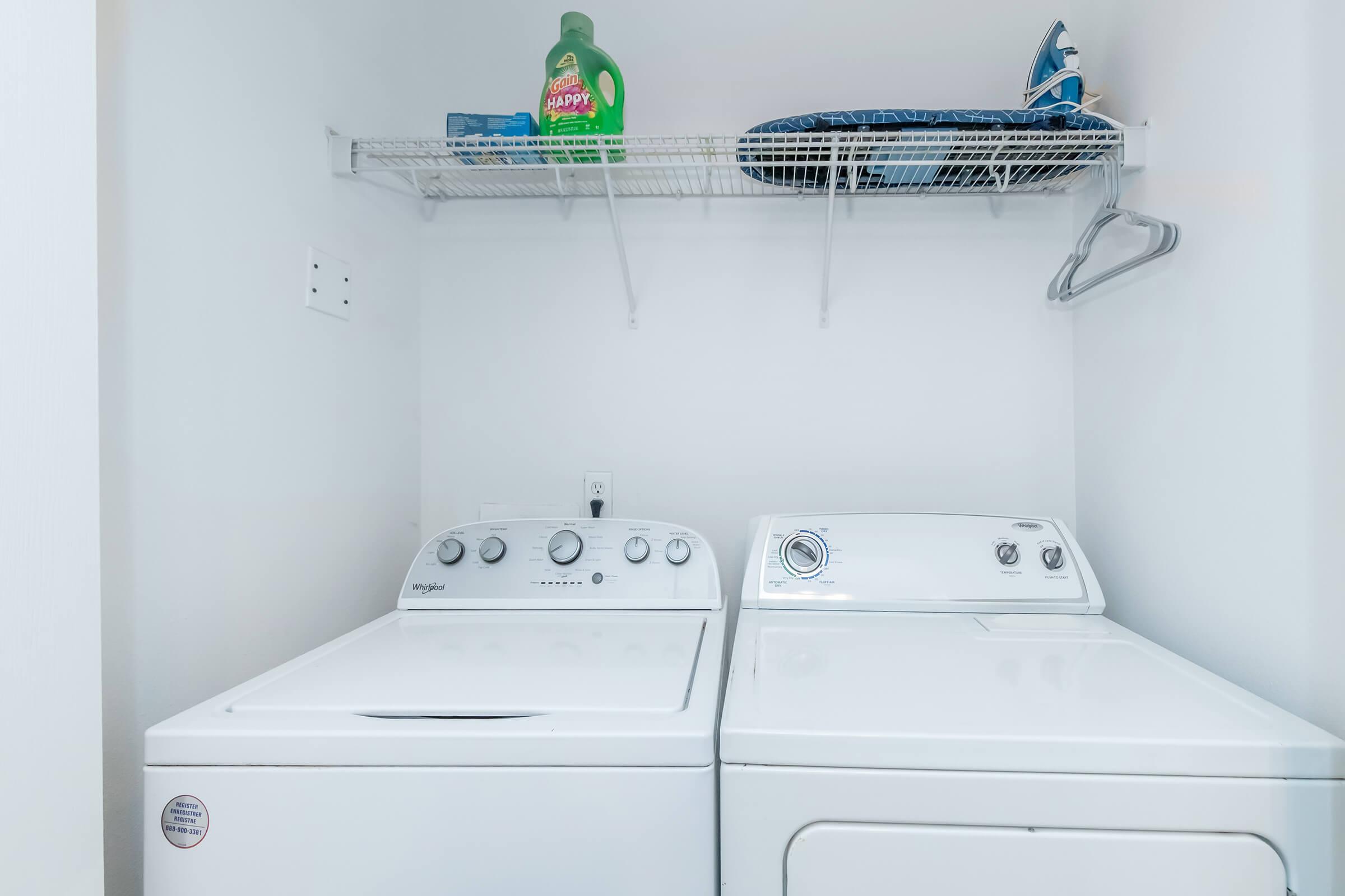 A clean laundry room featuring a white washing machine and dryer side by side. Above them, there is a shelf with a bottle of laundry detergent and an iron on an ironing board. The walls are light-colored, creating a simple and organized space.