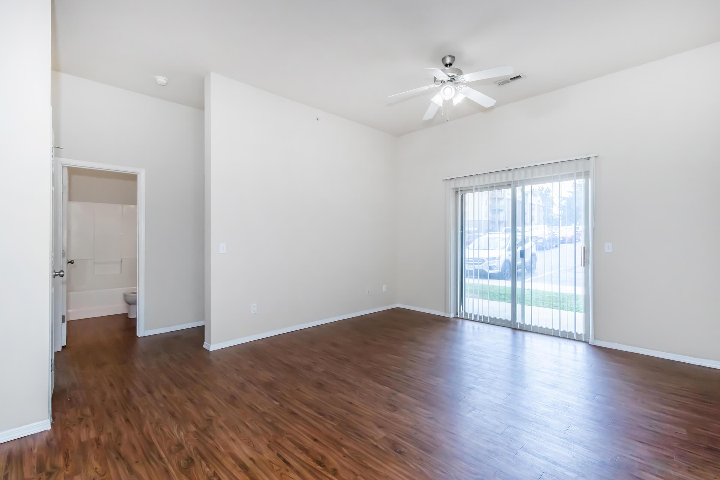 Spacious, well-lit living room with a ceiling fan and large windows showcasing sliding glass doors that lead to an outdoor area. The room features light-colored walls and hardwood-style flooring. A doorway on the left leads to a bathroom.