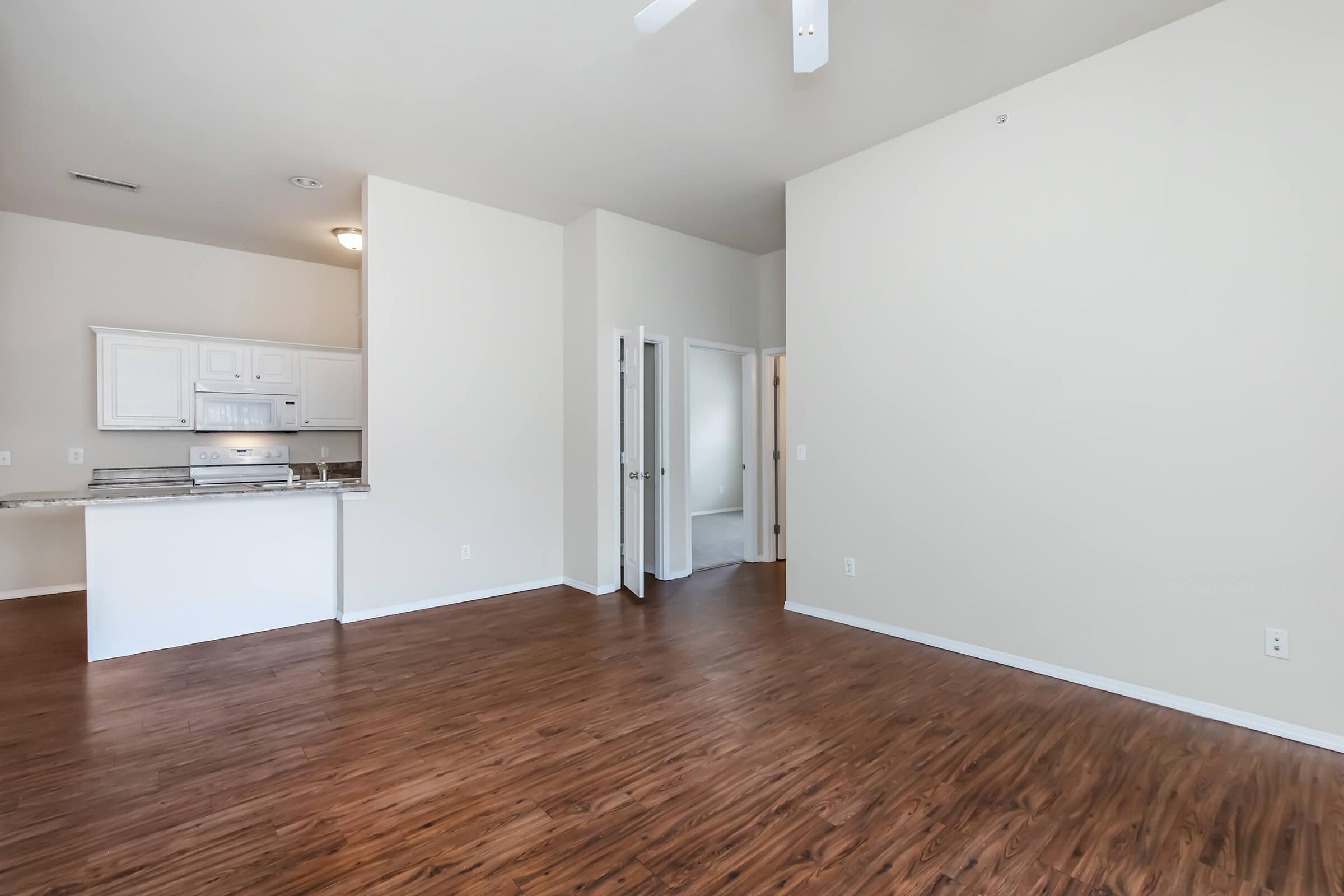 Spacious interior view of a modern apartment with wooden flooring, light-colored walls, and a ceiling fan. The room features an open layout, with a kitchenette visible in the background. There are doorways leading to other areas of the apartment, creating an inviting and airy atmosphere.