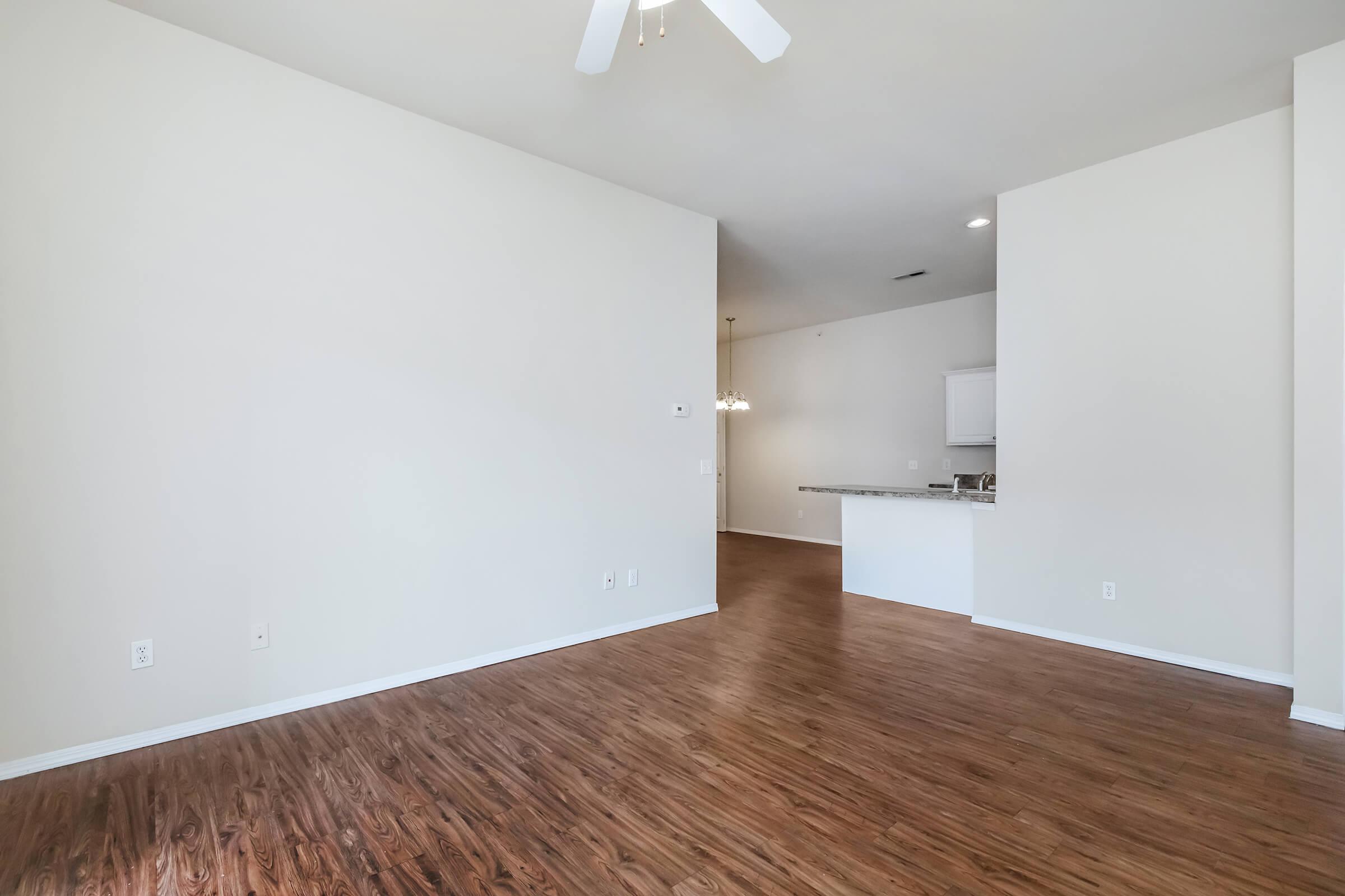 A spacious, empty living area with wooden flooring and light-colored walls. A ceiling fan is visible, and an open kitchen area with white cabinetry can be seen in the background. The space is well-lit and features no furniture, giving it a clean and airy feel.