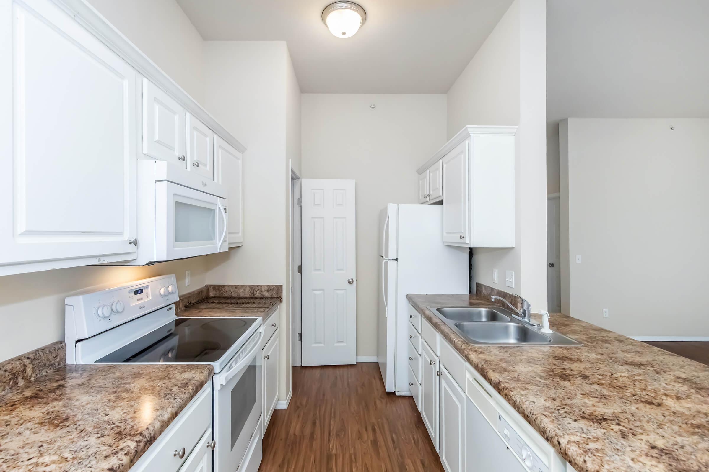 A modern kitchen featuring white cabinetry, stainless steel appliances, and granite countertops. There is an oven, microwave, and sink. The room has a light-colored wall and hardwood flooring, with a door leading to another room. Bright lighting from a ceiling fixture illuminates the space.