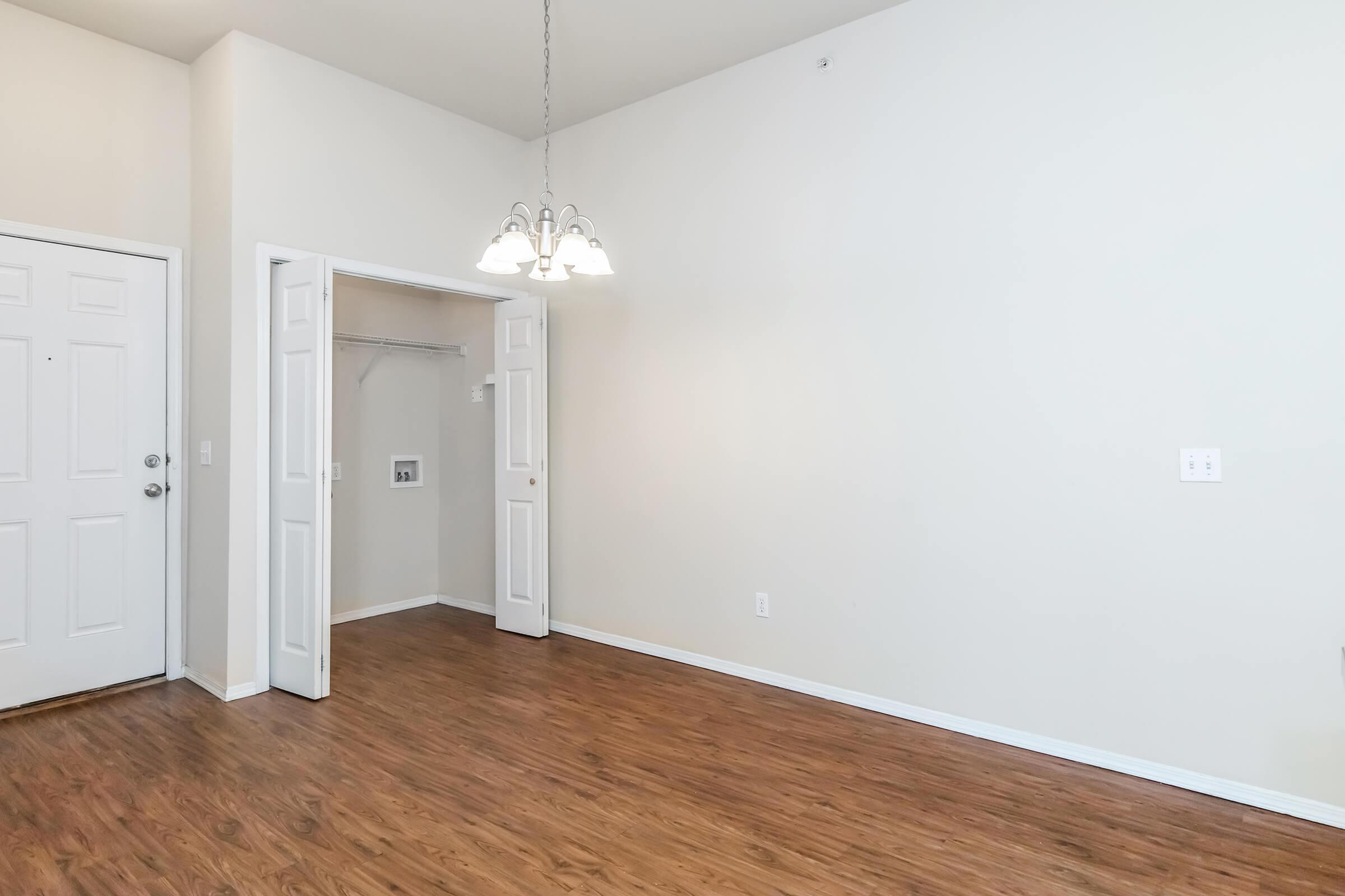 A spacious room with light-colored walls and a high ceiling. There is a wooden floor and a white door leading to a closet. A modern chandelier hangs from the ceiling. The entrance door is visible on the left side of the image, accentuating the open and airy feel of the space.