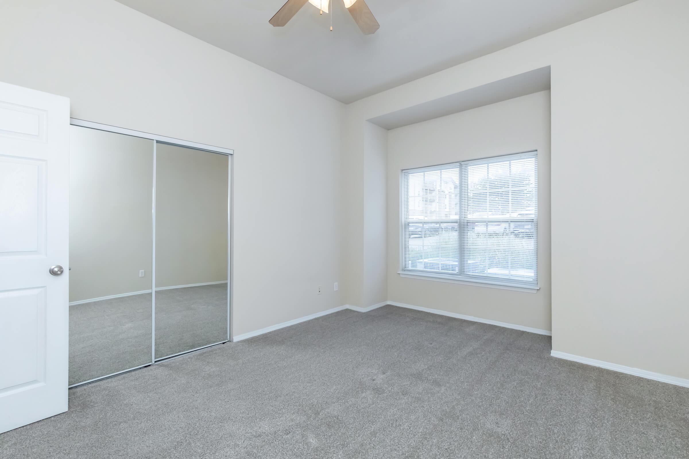 A spacious, empty bedroom featuring light-colored walls and a window with blinds. There's a ceiling fan and a sliding mirror closet door on one wall. The floor is covered with light gray carpet, creating a clean and airy ambiance. The room has natural light, enhancing its openness.