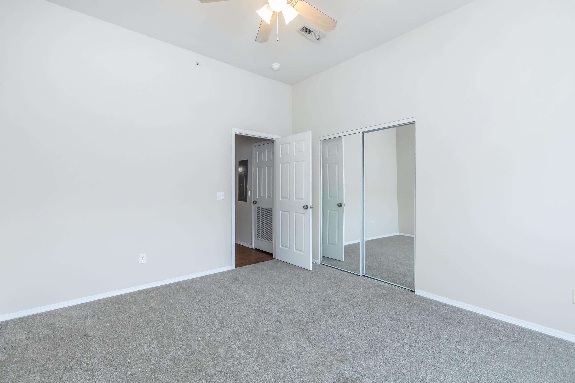 A spacious room with light beige walls and carpet flooring. There is a ceiling fan, a closet with mirrored sliding doors, and an open doorway leading to another area. Natural light brightens the space, giving it an inviting atmosphere.