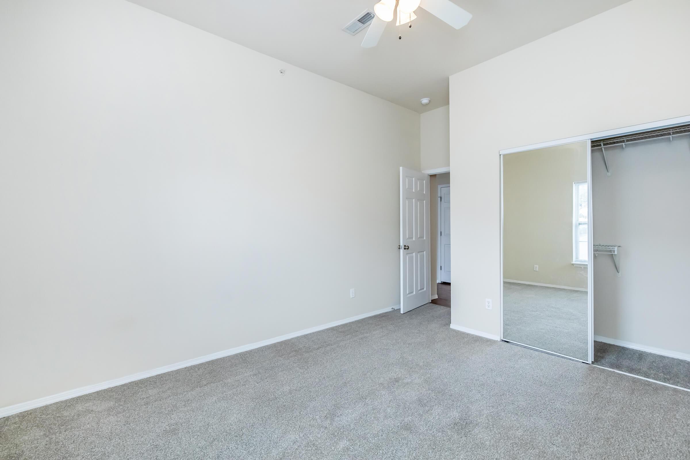 A spacious, empty room with light-colored walls and carpet. The room features a ceiling fan, a mirrored closet door, and a doorway leading to another area. Natural light enters through a nearby window. The overall ambiance is bright and open, suitable for various purposes.