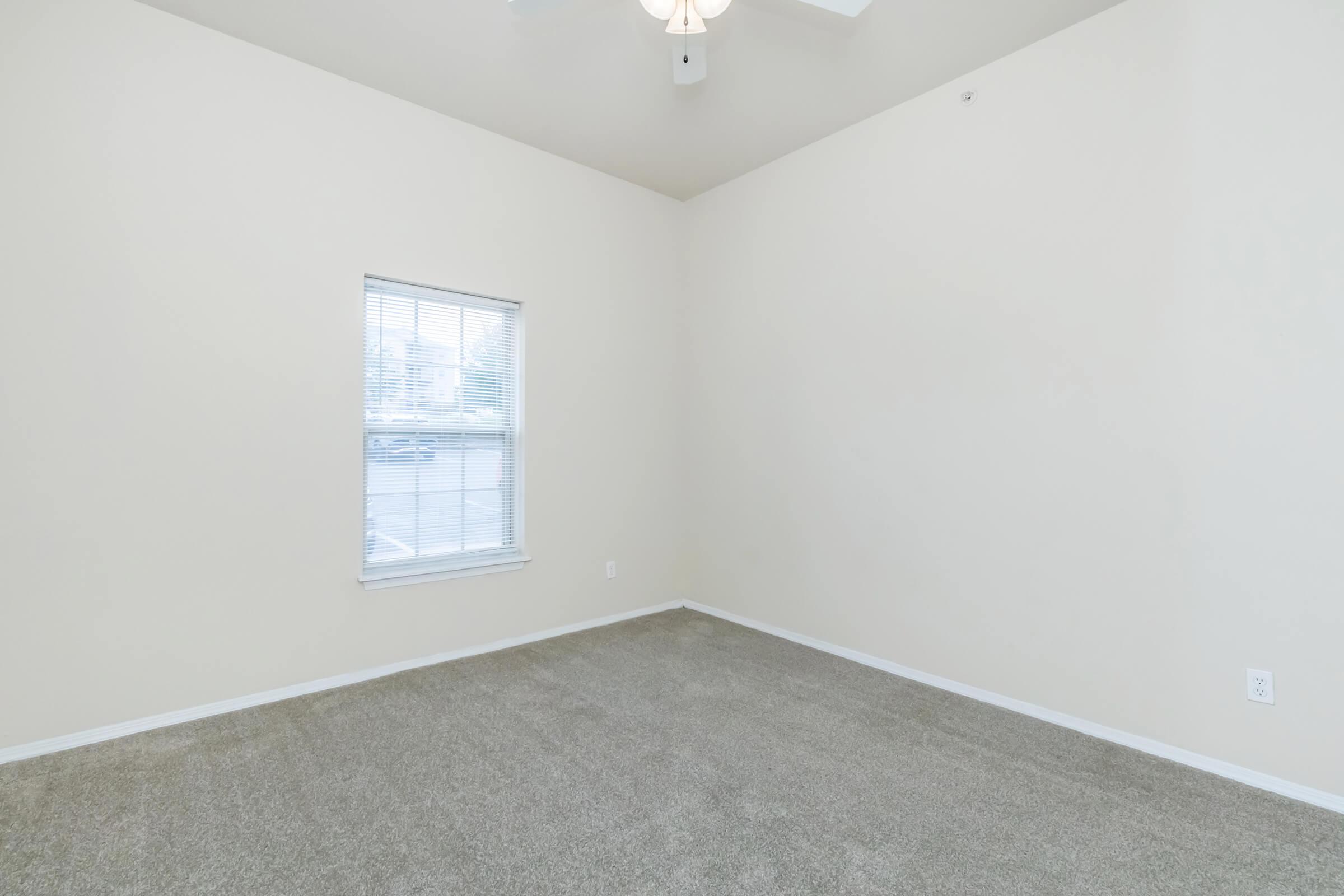 Empty room with light beige walls and a light gray carpet. A single window with white blinds allows natural light to enter. The ceiling features a ceiling fan, and there is minimal furniture or decor, creating a spacious feel.