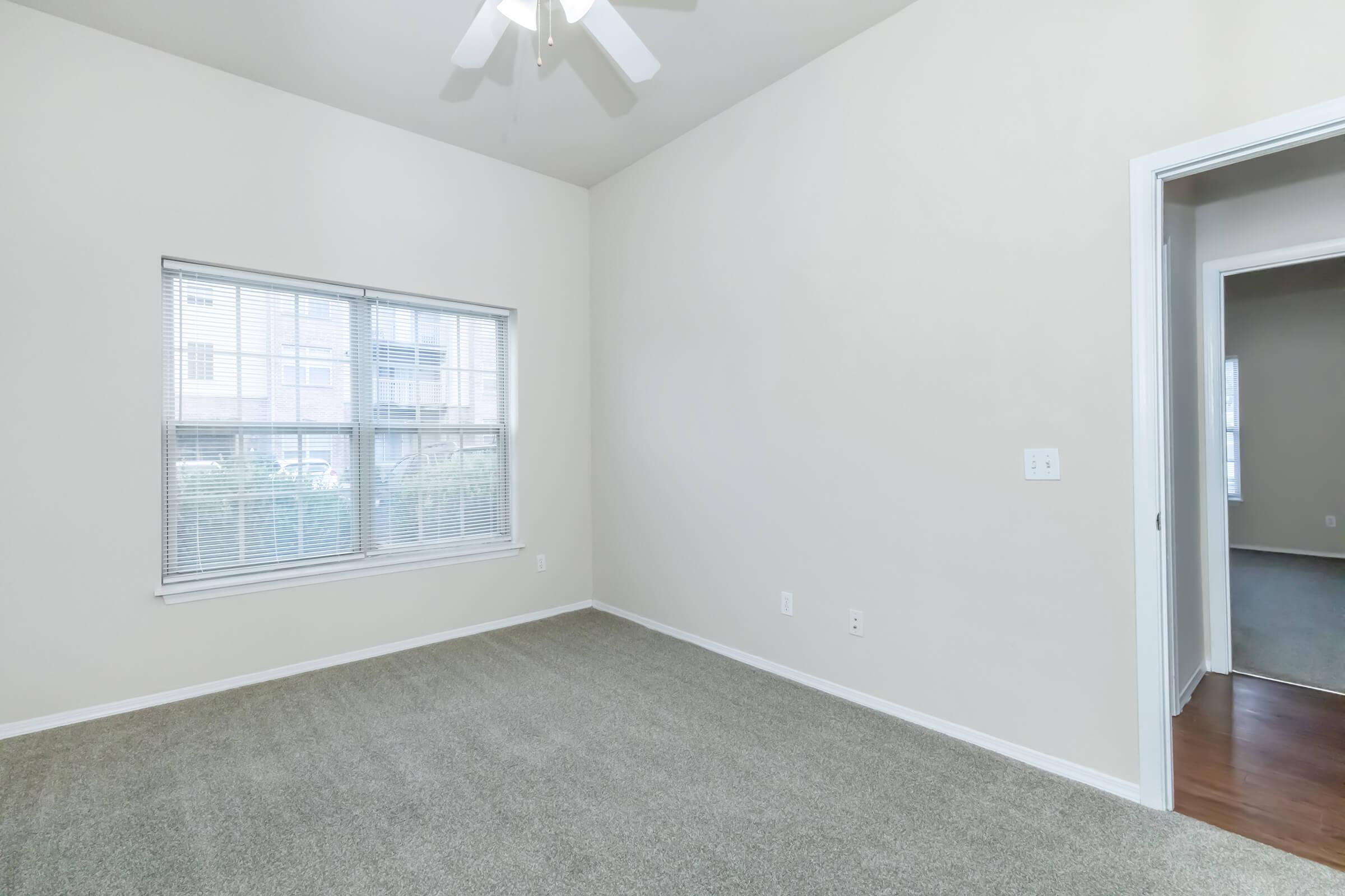 A spacious, well-lit room with light-colored walls and a ceiling fan. The flooring is carpeted in a neutral shade. A large window allows natural light to enter, and a doorway leads to another room, suggesting potential for versatile use of the space.