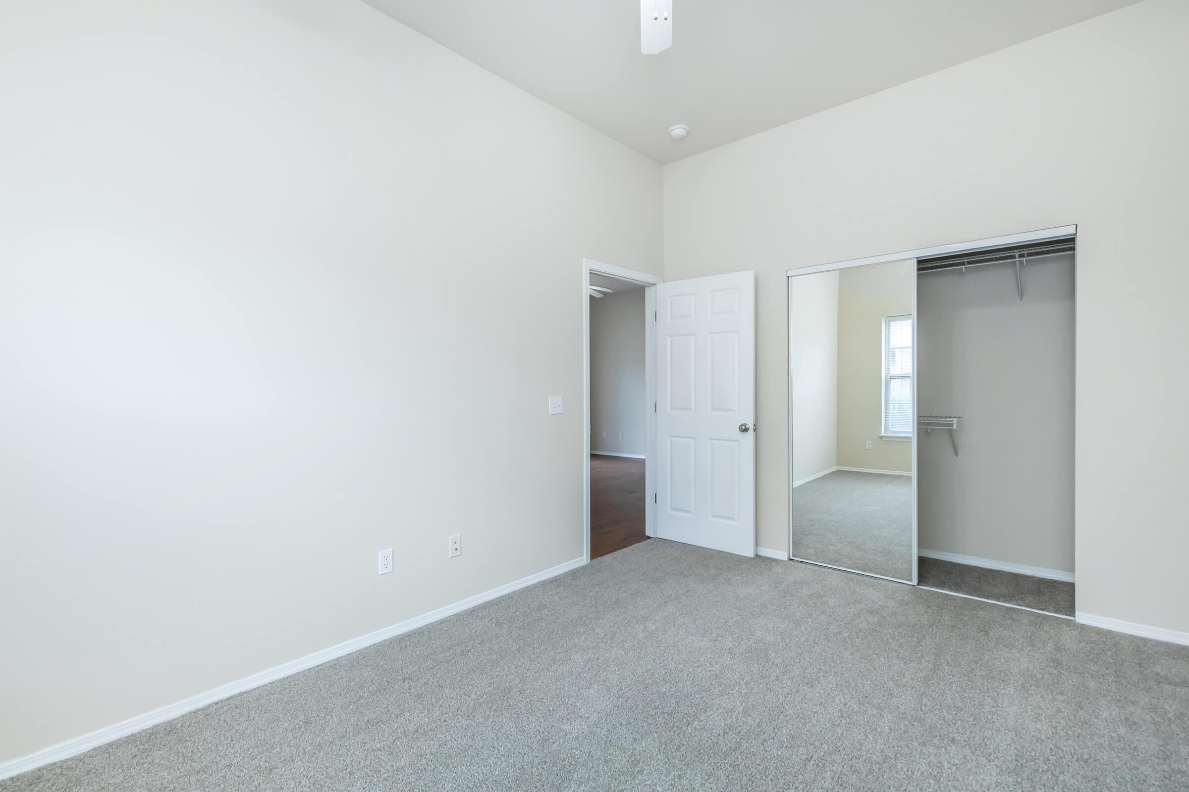A spacious, empty room featuring beige walls and a high ceiling. The floor is covered in light gray carpeting. A white door leads to another room, while a mirrored closet door reflects the interior. Natural light streams in through a window, creating a bright and airy atmosphere.