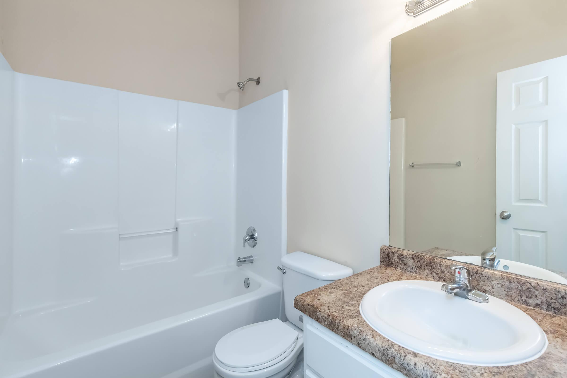 A clean, modern bathroom featuring a white bathtub with a showerhead, a sink with a round basin, and a mirror above the sink. The countertop is light brown with speckles, and there is a toilet nearby. The walls are painted a soft beige, and a towel rack is visible on the wall opposite the mirror.
