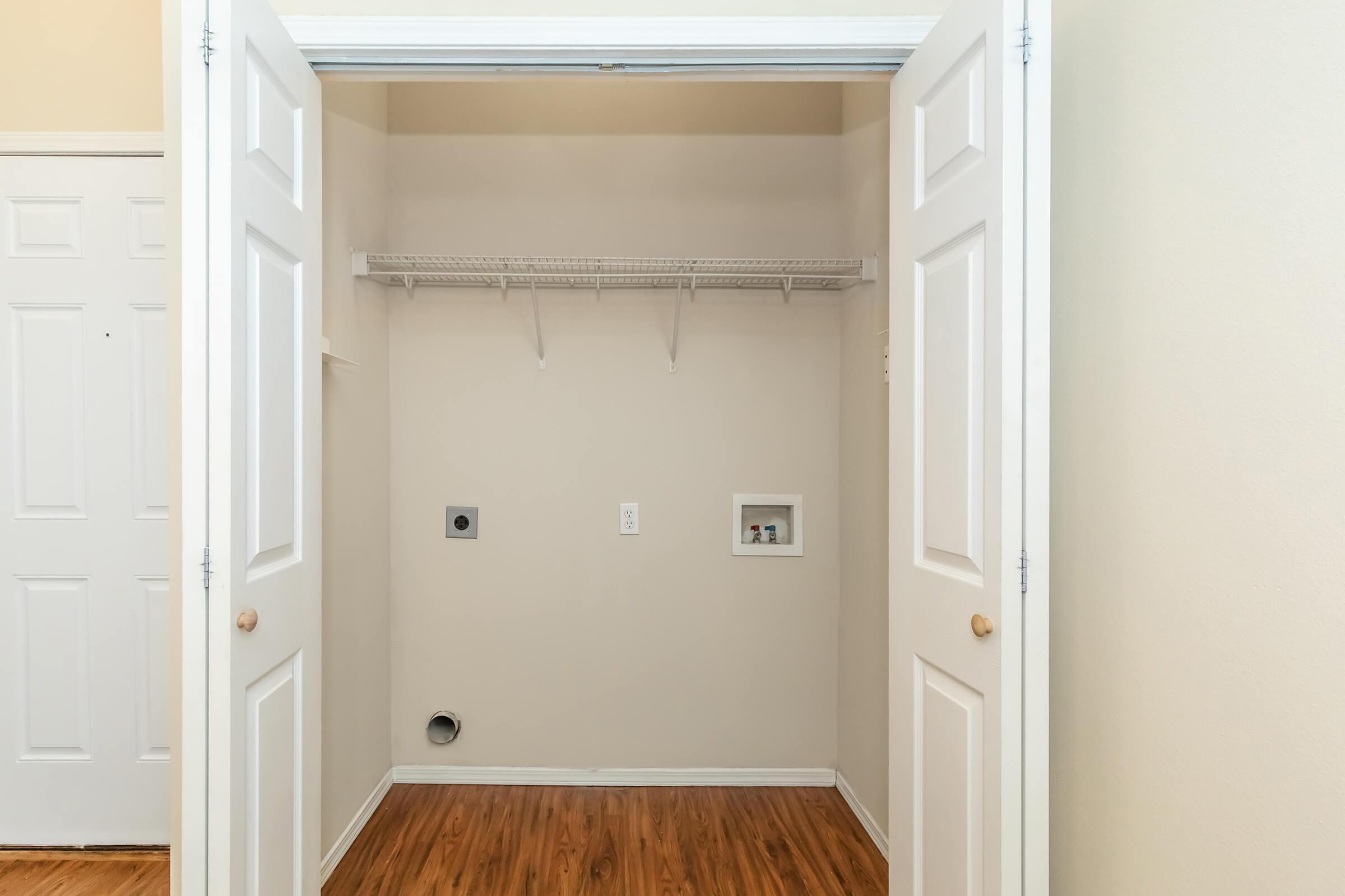 An empty closet space with white walls and brown hardwood flooring. The closet has two white double doors, a shelf along the top, and two wall-mounted electrical outlets. There is a small opening on the wall for a vent or plumbing. The area appears clean and well-maintained.