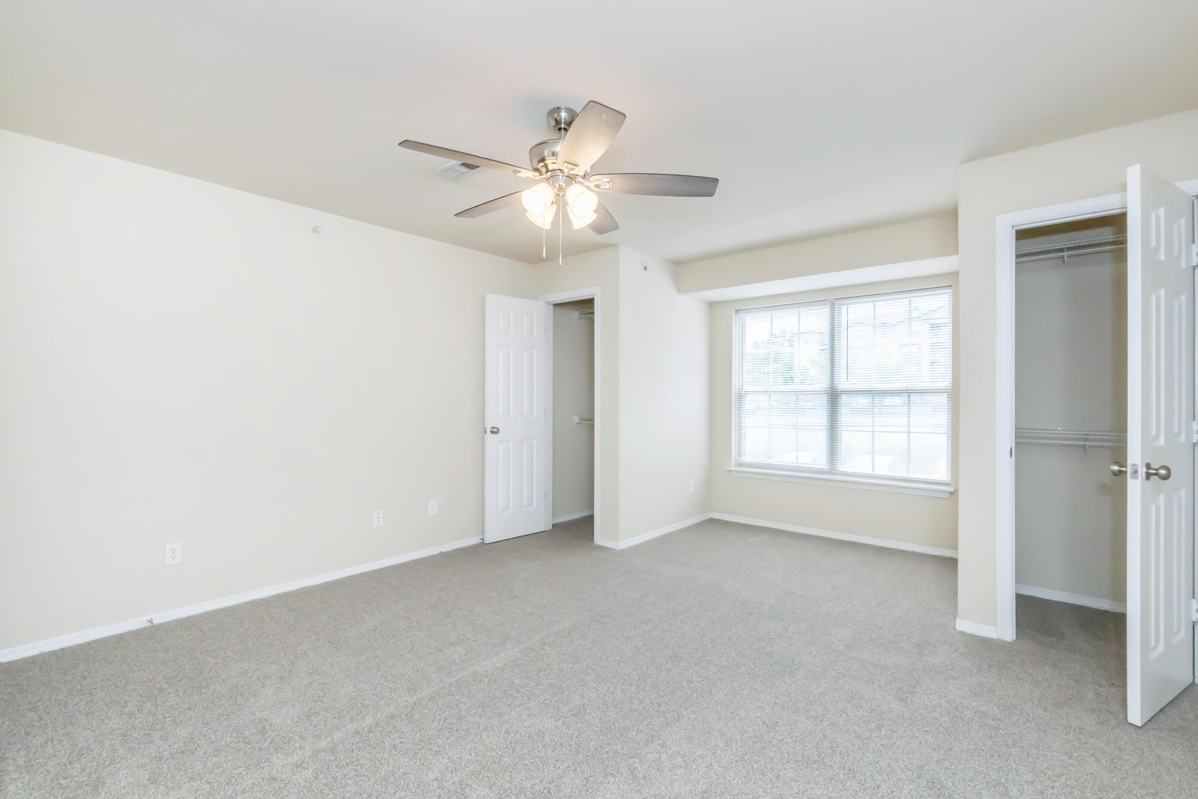 A spacious, empty room with light beige walls and carpet flooring. There's a ceiling fan, a window allowing natural light in, and an open closet with sliding doors. Two doors are visible; one leading to another space and the other possibly a bathroom or closet.