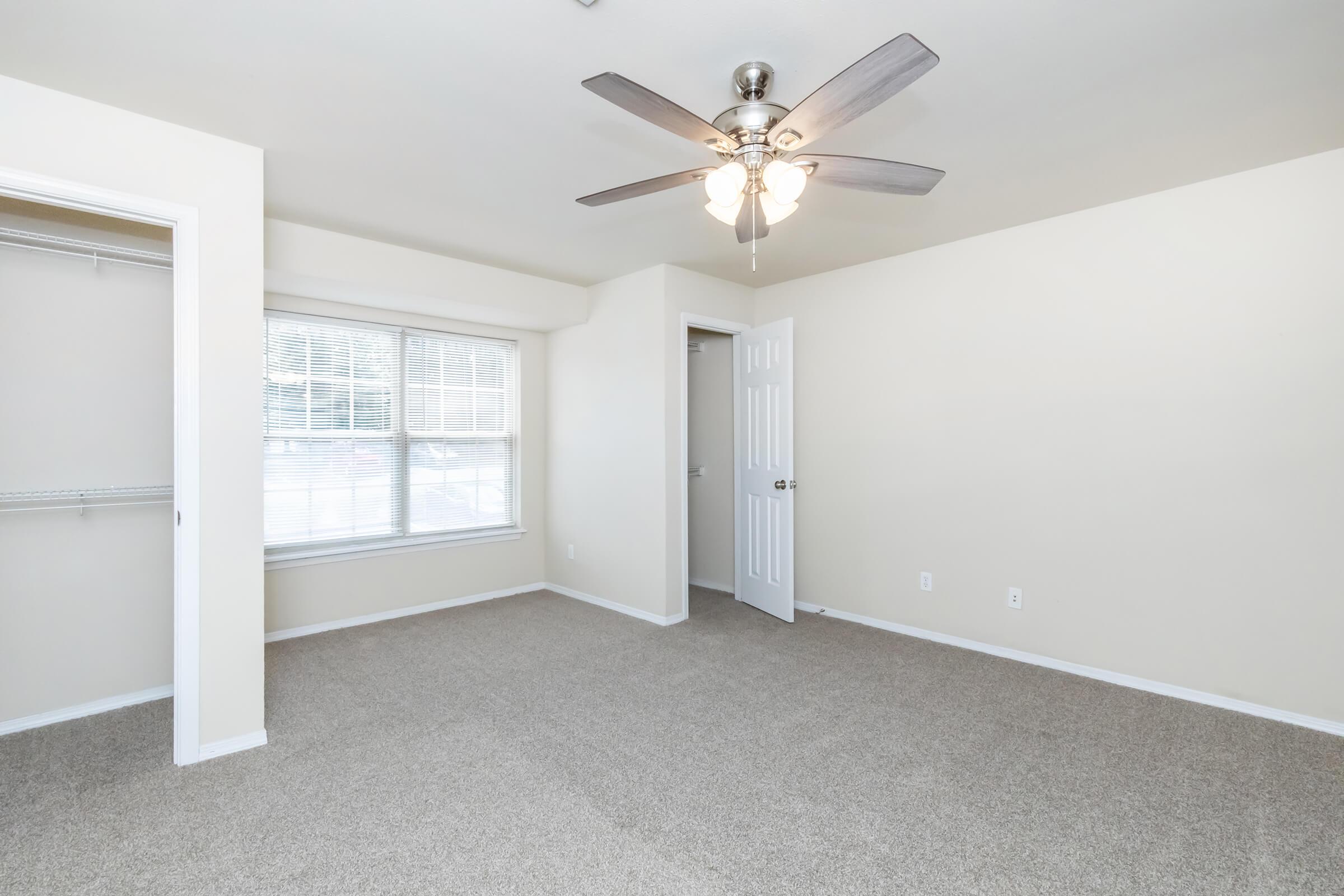 A spacious, well-lit room with beige walls and a light-colored carpet. It features a ceiling fan with four lights, a large window with blinds, and a closet with an open door. The room has a simple, clean design, making it suitable for various setups.