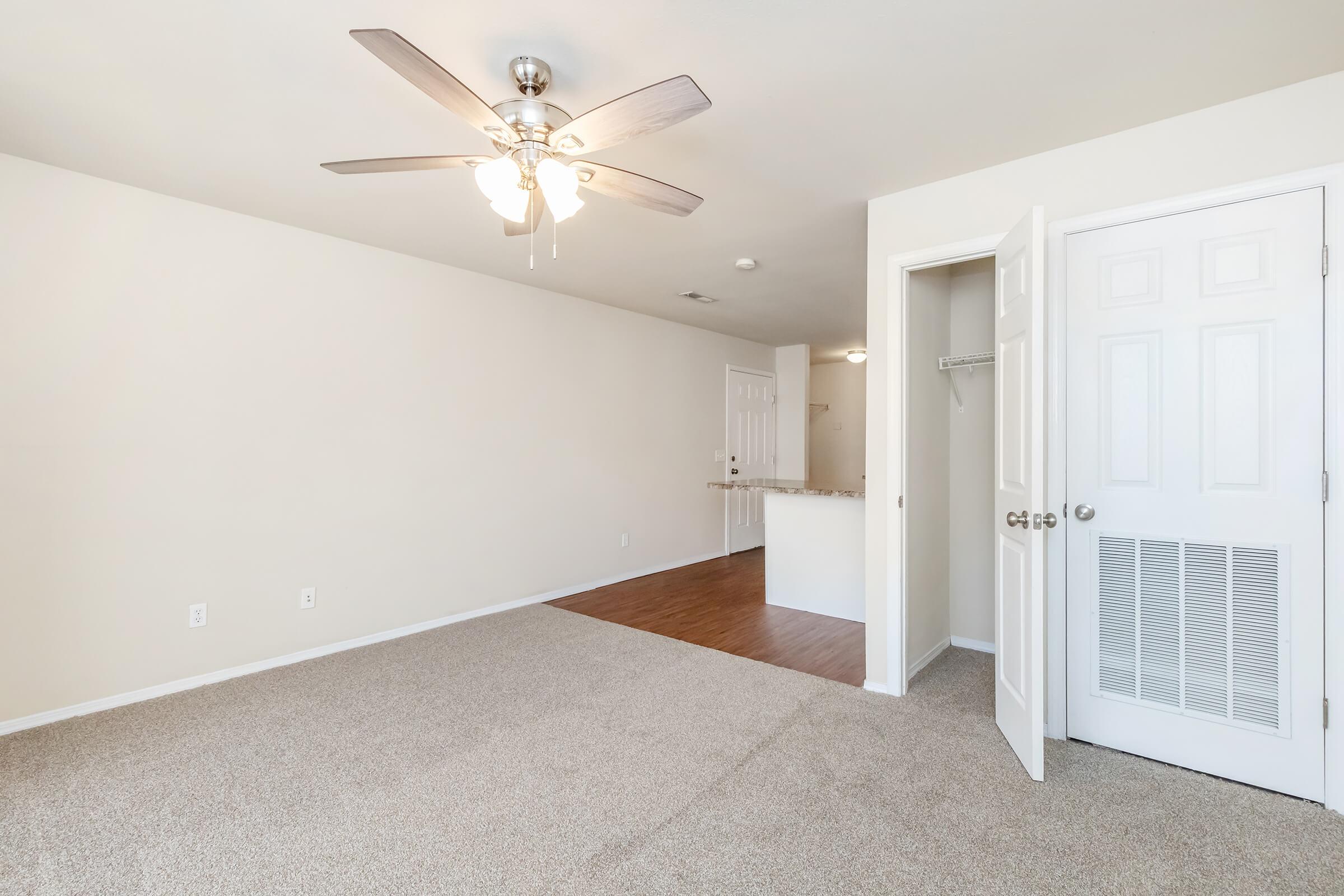 A spacious room with beige walls and carpeted flooring. It features a ceiling fan, a closed door leading to another room, and a doorway in the background. The room has a light and airy feel, with a small area that leads to a kitchen or dining space.