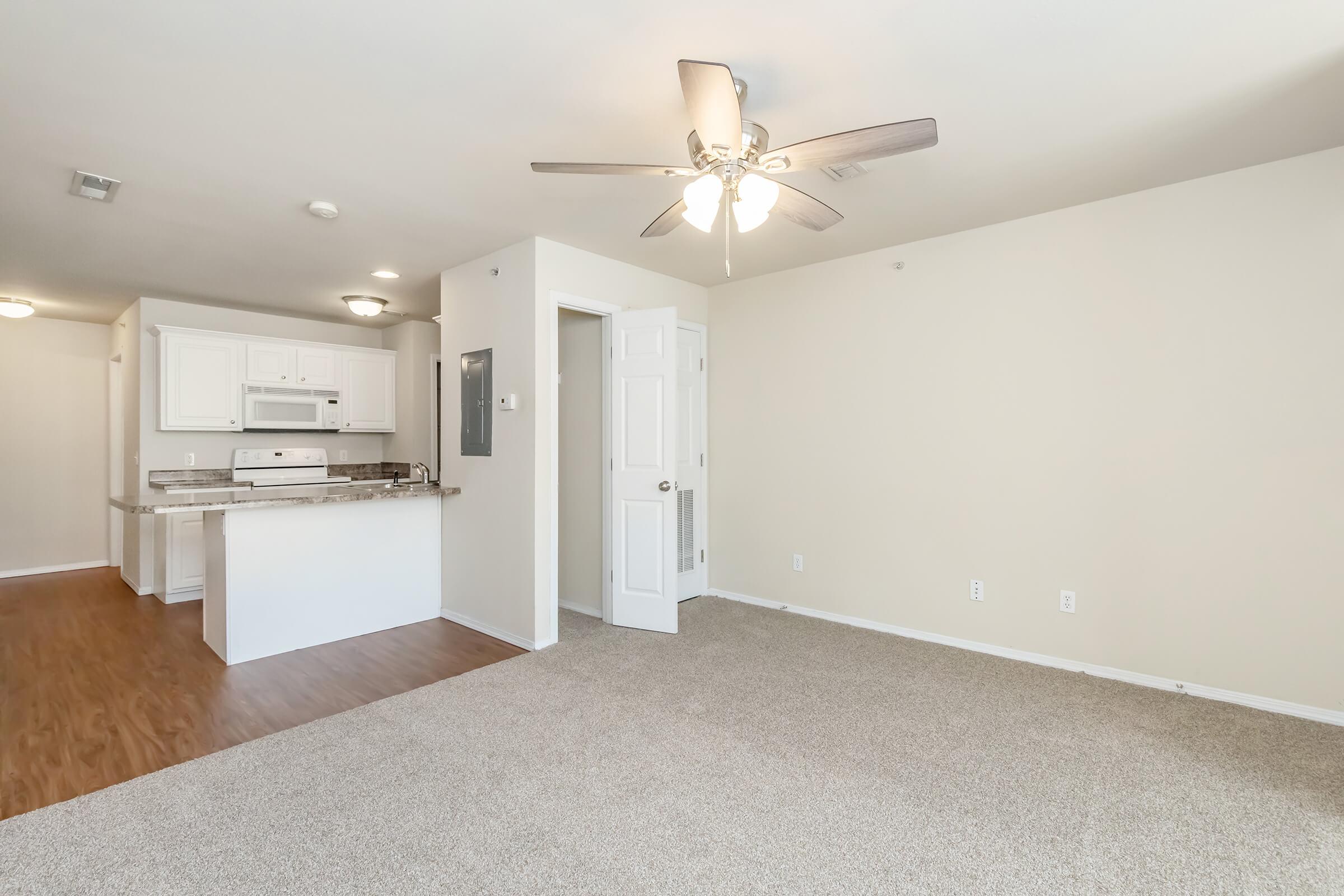 Spacious living area with light-colored walls and a ceiling fan. Features beige carpeting and laminate flooring. A small kitchen area with white appliances and cabinets is visible in the background, along with a doorway leading to another room. Natural light fills the space.