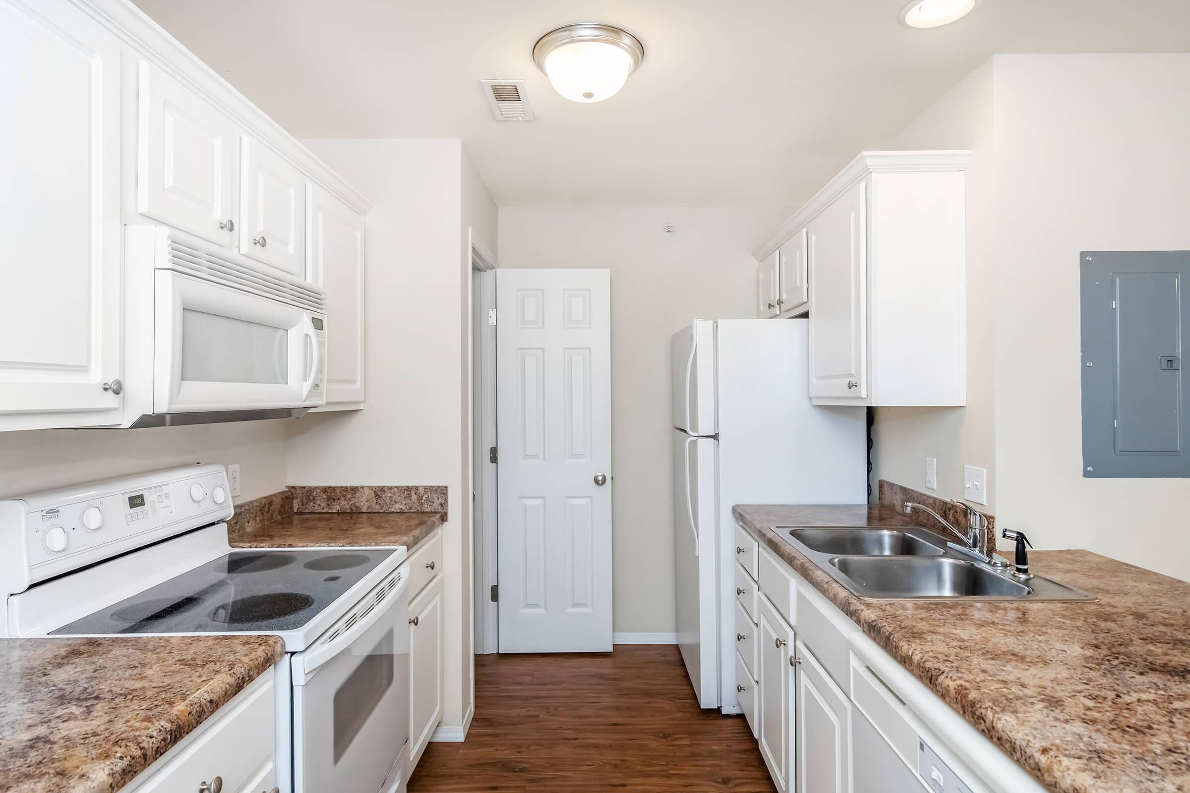 A modern kitchen featuring white cabinetry, granite countertops, and stainless steel appliances. Includes an oven, microwave, refrigerator, and dual sinks, with a door leading to another room and a wall-mounted electrical panel. The floor is composed of wood-like laminate.