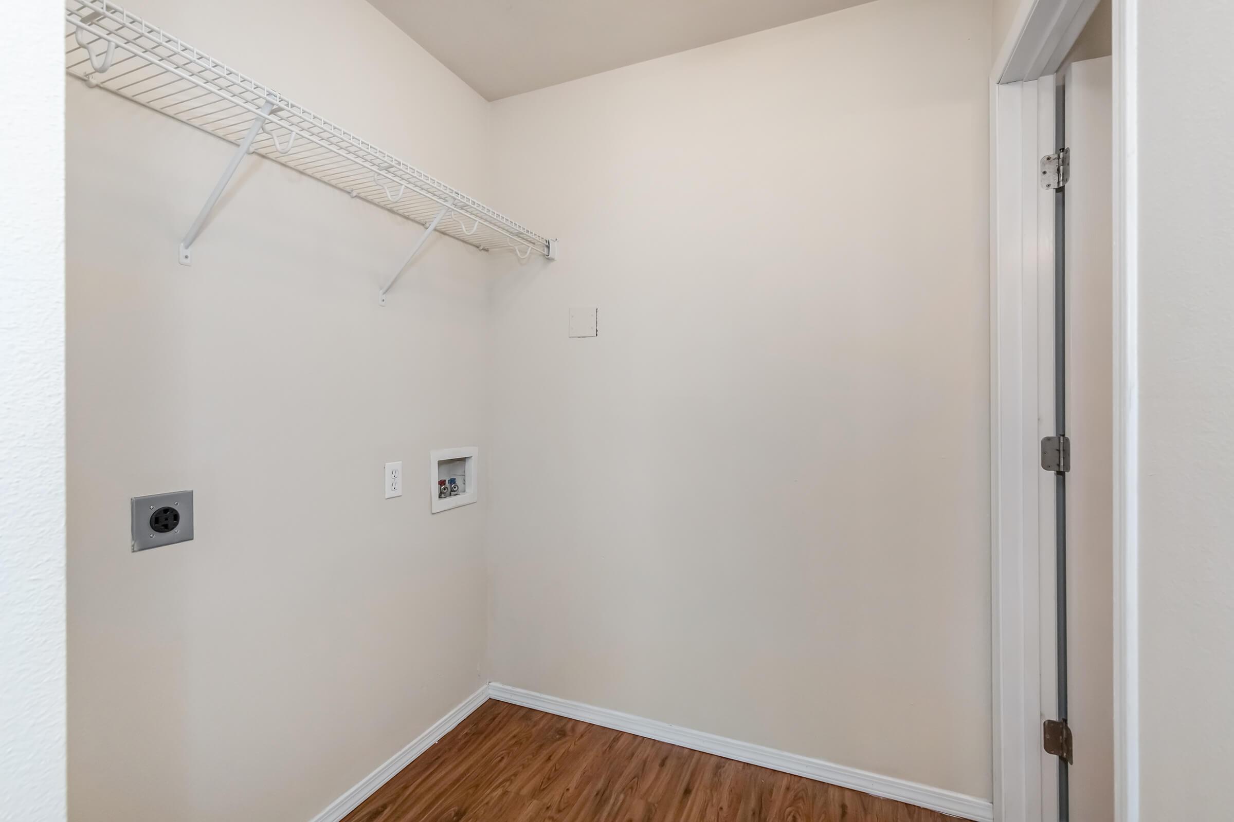A small, empty room with light-colored walls and a wooden floor. The space features a metal wire shelf on one side and two electrical outlets on the wall. A door partially visible on the right suggests entrance or exit points. The overall appearance is clean and uncluttered.