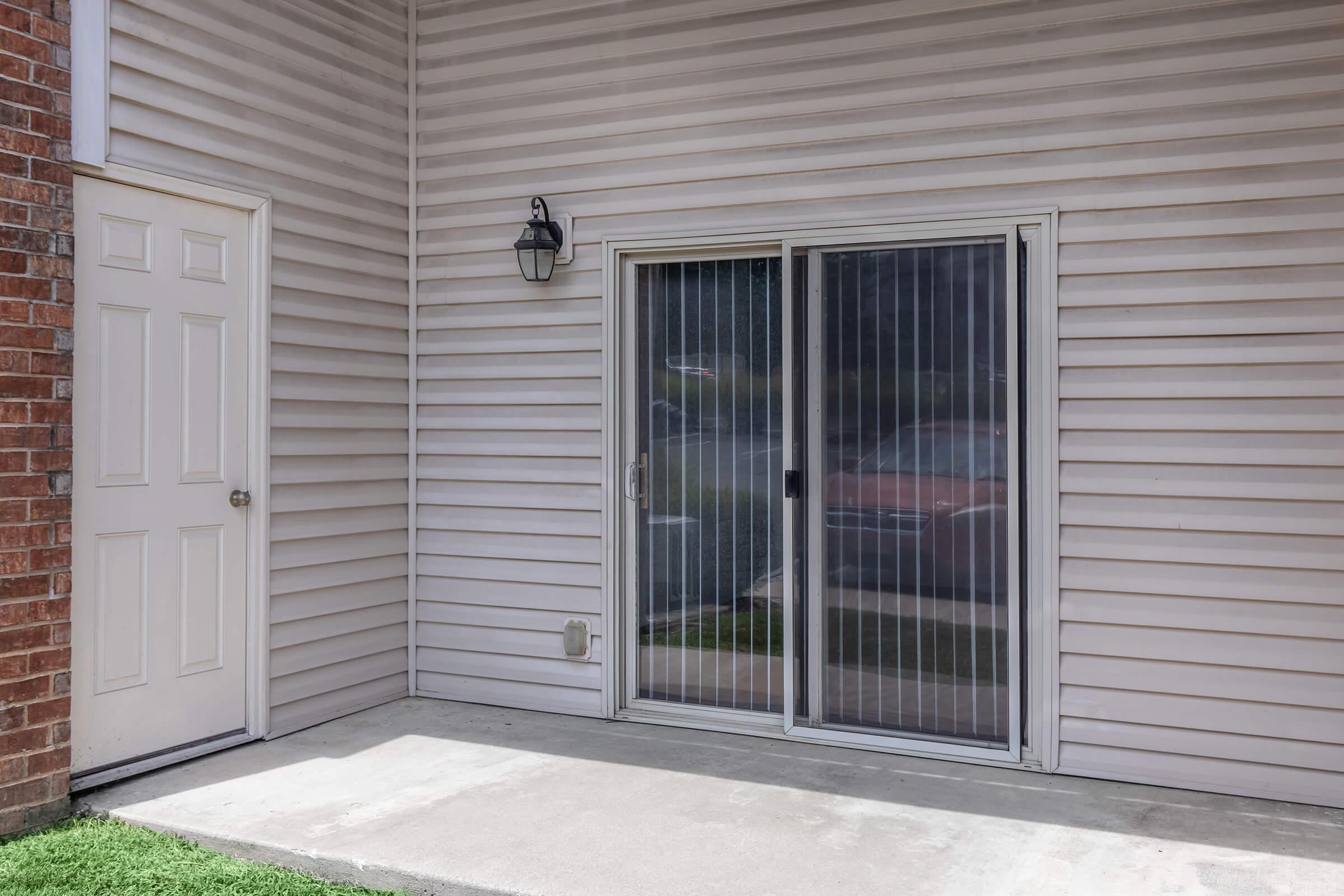 A small exterior patio area featuring a sliding glass door with vertical blinds, next to a white door. The wall has light-colored siding, and there is a patch of green grass in front of the patio. The setting appears to be part of a residential building.
