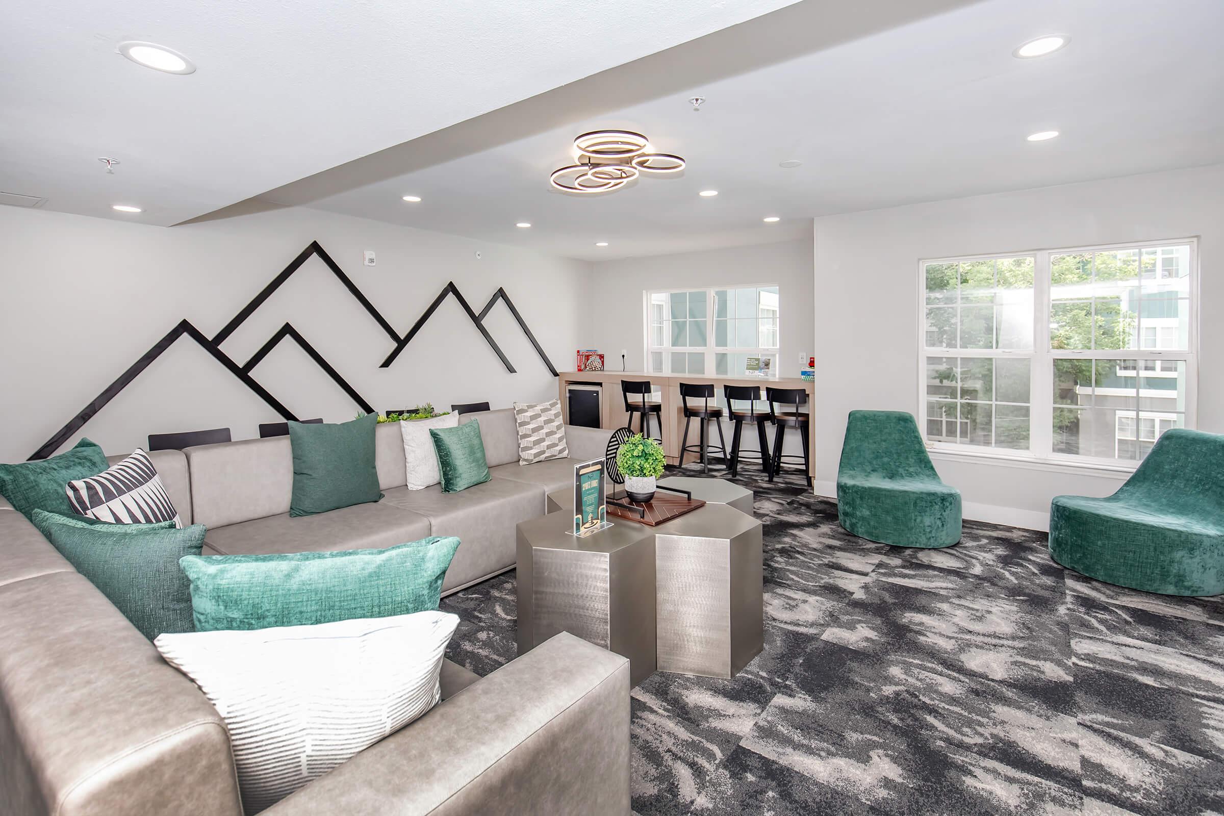 A modern indoor lounge area featuring a large grey sectional sofa with colorful accent pillows, sleek metal coffee tables, and distinct green seating. The walls are decorated with geometric patterns, and large windows provide ample natural light. A bar-style table with stools is visible in the background.