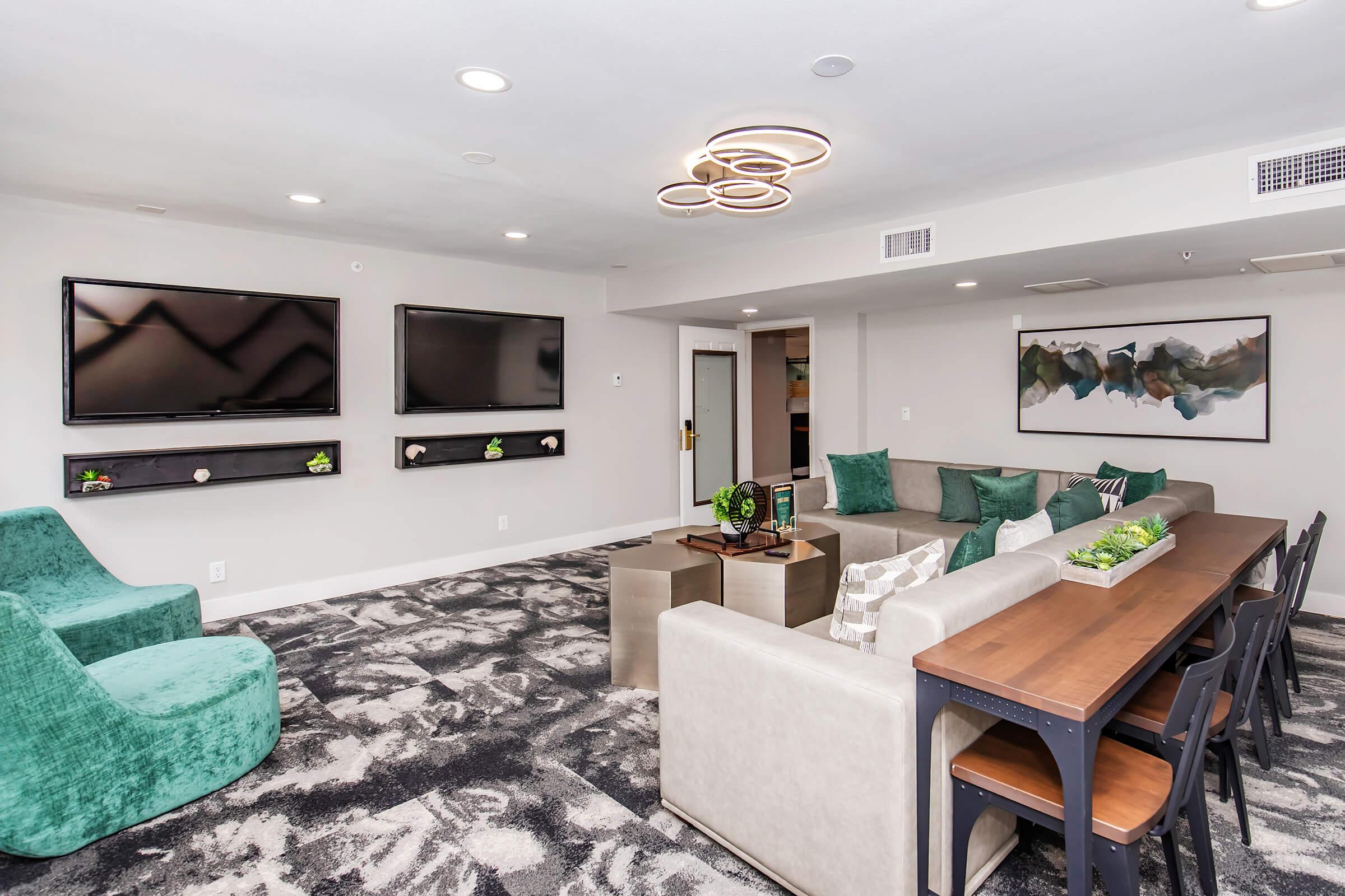 A modern lounge area featuring two large flat-screen TVs mounted on a wall, a cozy seating arrangement with green armchairs and a light-colored sofa, a wooden dining table with black chairs, and decorative plants. The space has a contemporary design with stylish lighting and neutral colors.