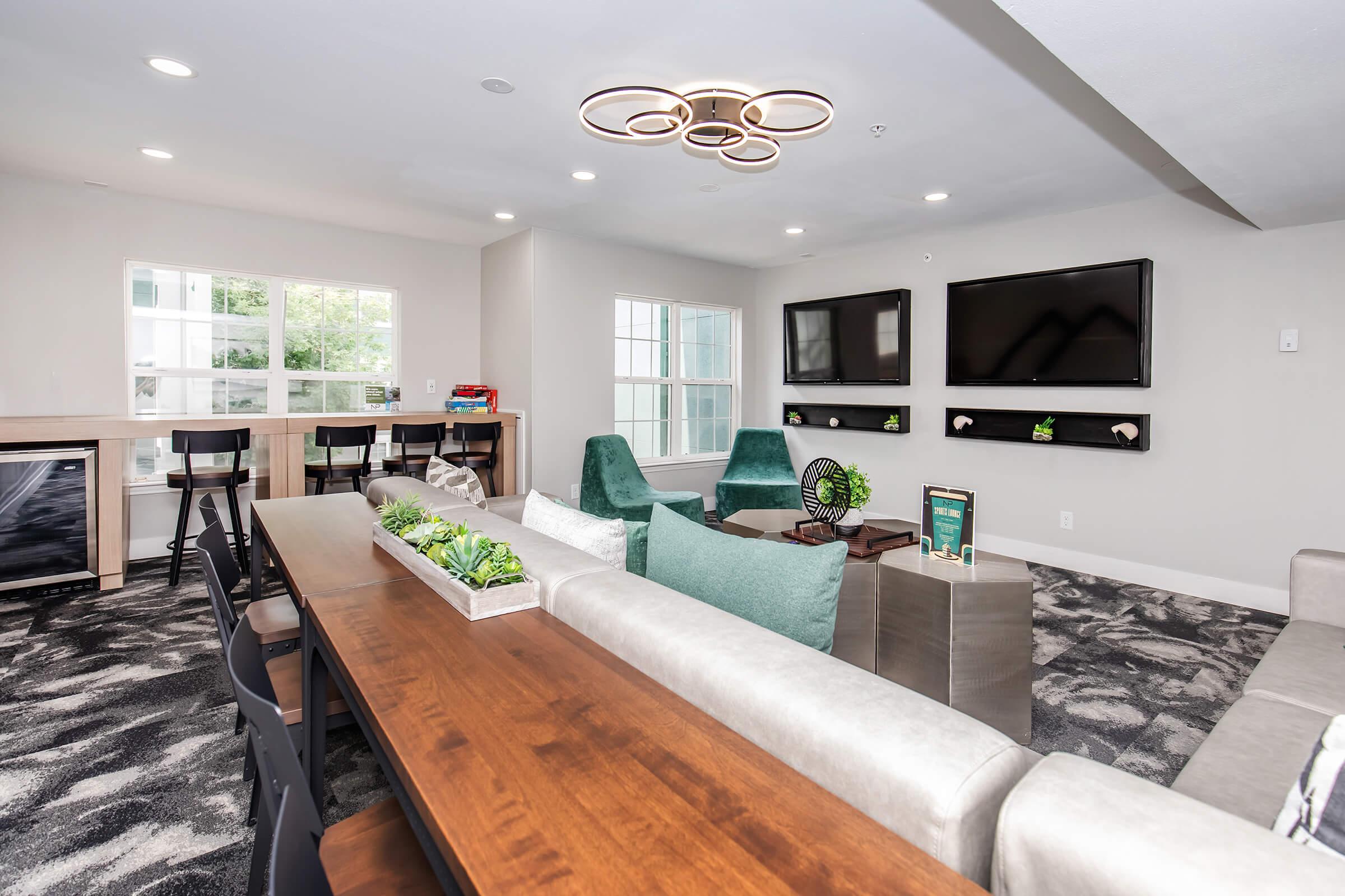 Modern lounge area featuring comfortable seating with green accents, a wooden dining table, and wall-mounted flat-screen TVs. Large windows allow natural light, and decorative plants add a touch of nature. A fan and various books are visible on a shelf, creating a welcoming atmosphere for relaxation or socializing.