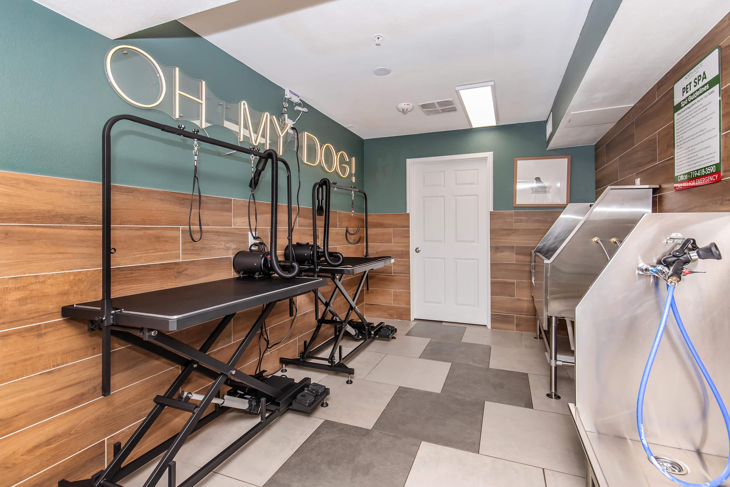 A pet grooming area featuring two grooming tables with adjustable arms, a door leading to another space, and a stainless steel bathing tub. The wall is decorated with wooden panels and a playful sign that reads "OH MY DOG!"
