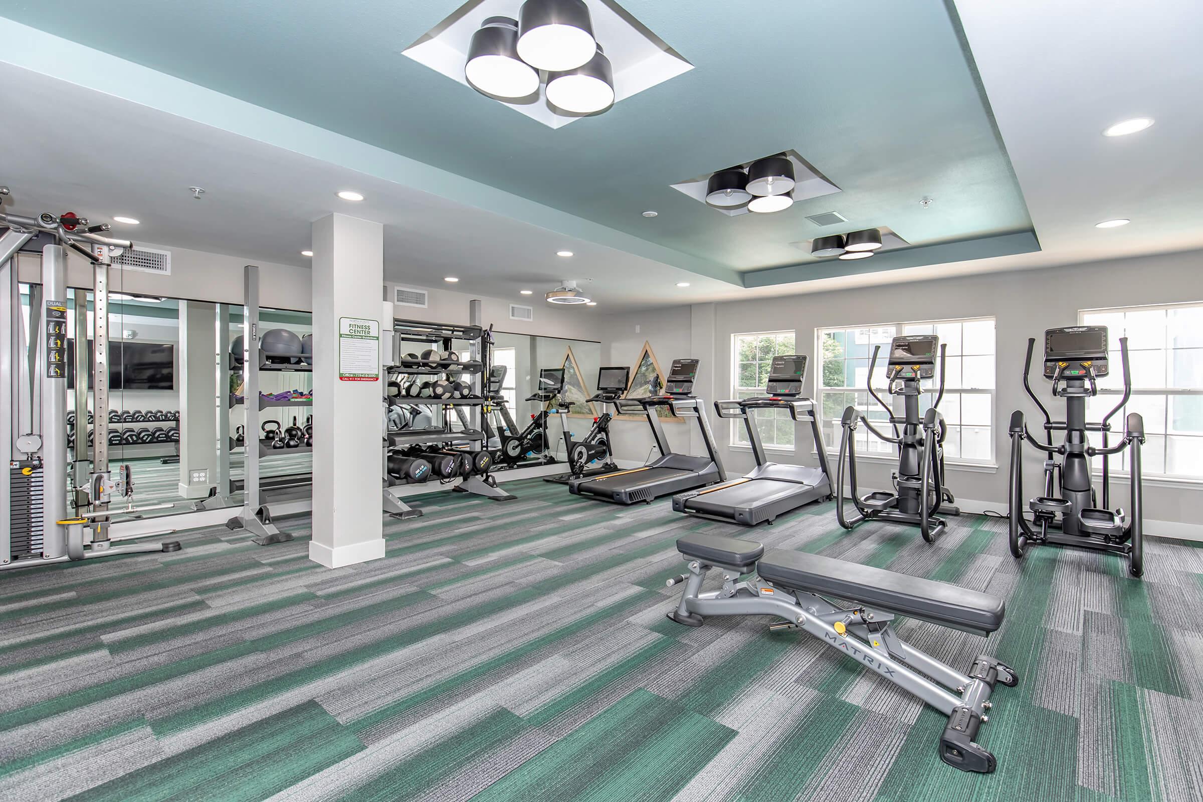 A modern gym featuring various exercise equipment, including treadmills, ellipticals, and free weights. The flooring has a colorful striped pattern, and large windows allow natural light to enter the space. The room is well-lit with overhead lighting and has a clean, open layout.