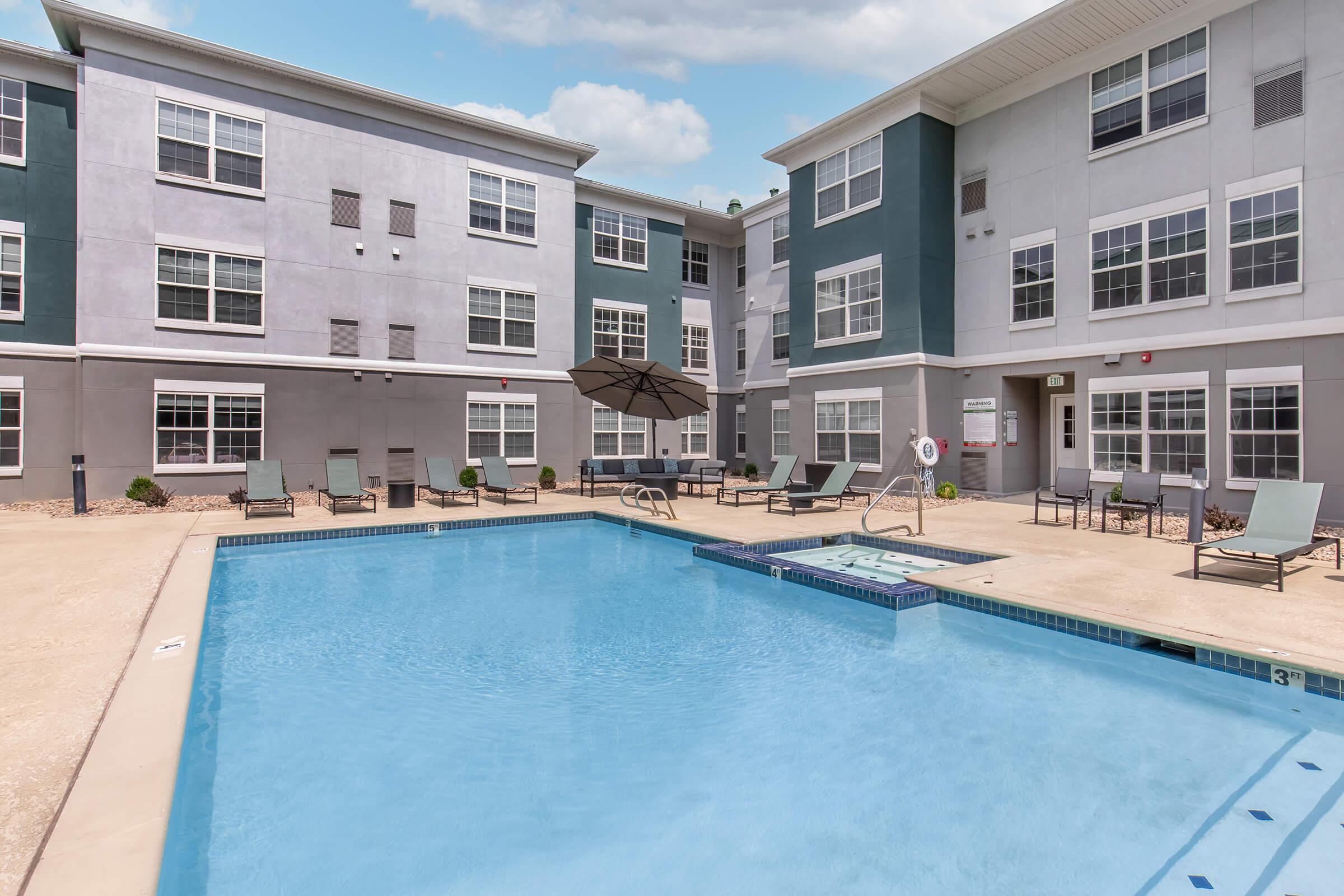 A clear swimming pool surrounded by lounge chairs and an umbrella, set within a courtyard of a multi-story apartment building. The building features a modern design with a mix of light and dark gray colors, large windows, and a bright sky overhead.