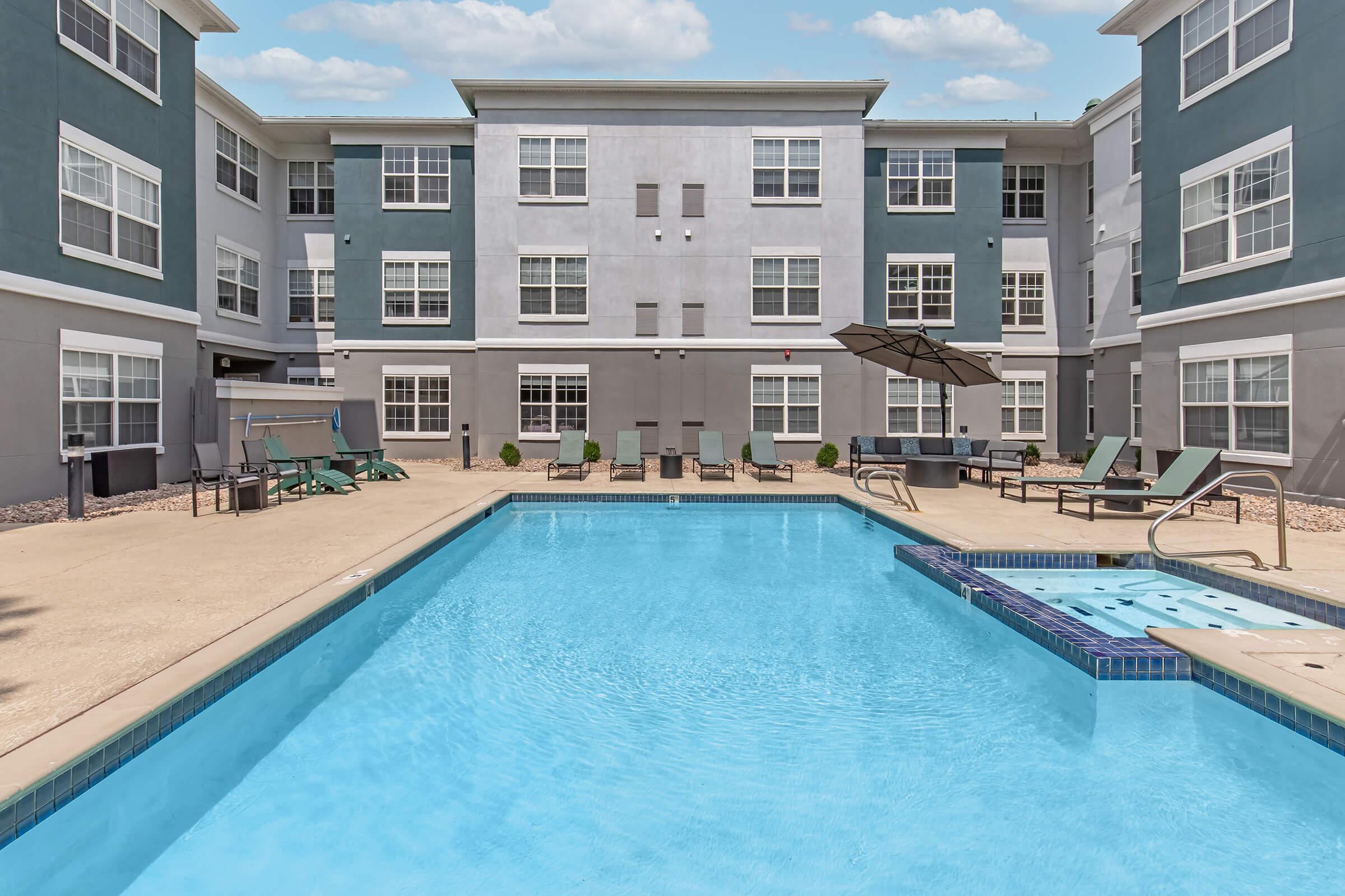 A bright, inviting outdoor pool area surrounded by lounge chairs and umbrellas, with a modern apartment building in the background. The pool is clear and blue, and the area is well-lit, creating a relaxing atmosphere for residents and guests.