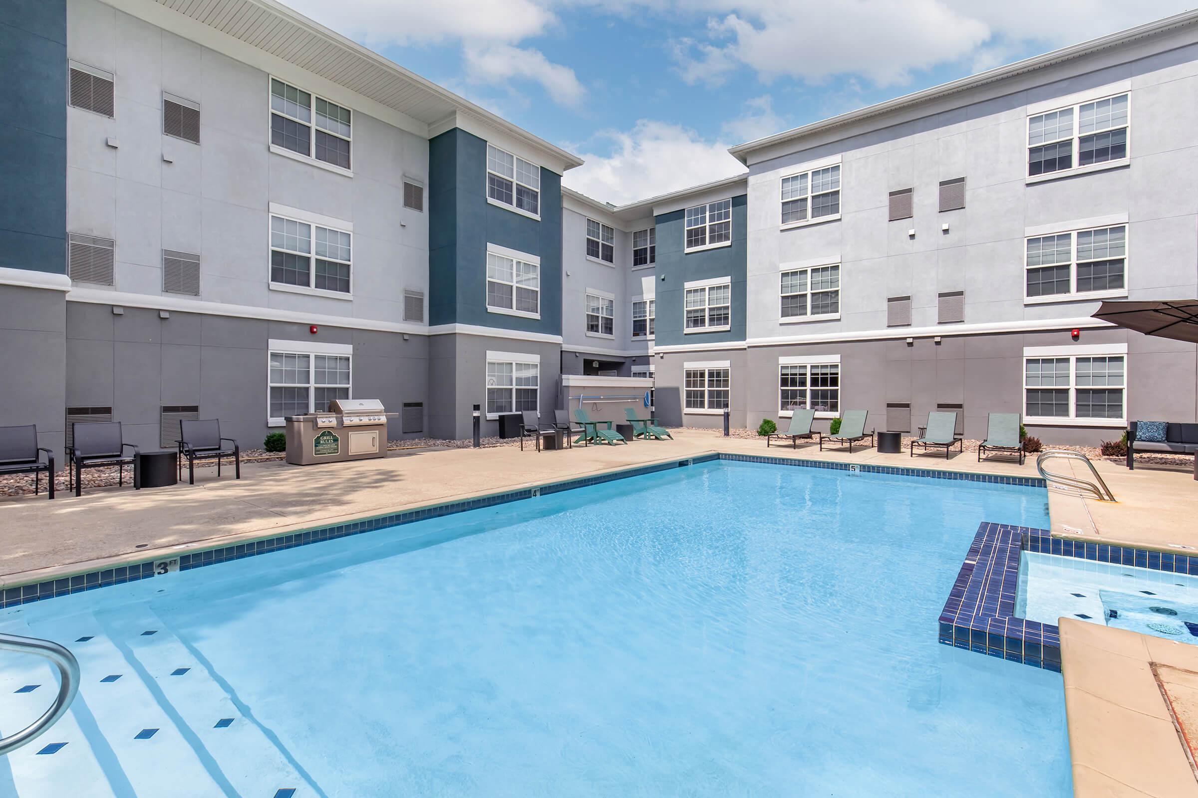 A clear swimming pool surrounded by lounge chairs and patio furniture, against a backdrop of a modern multi-story building with gray and blue exterior. The scene is bright and inviting, featuring a small hot tub next to the pool. The sky is partly cloudy, adding to the pleasant atmosphere.