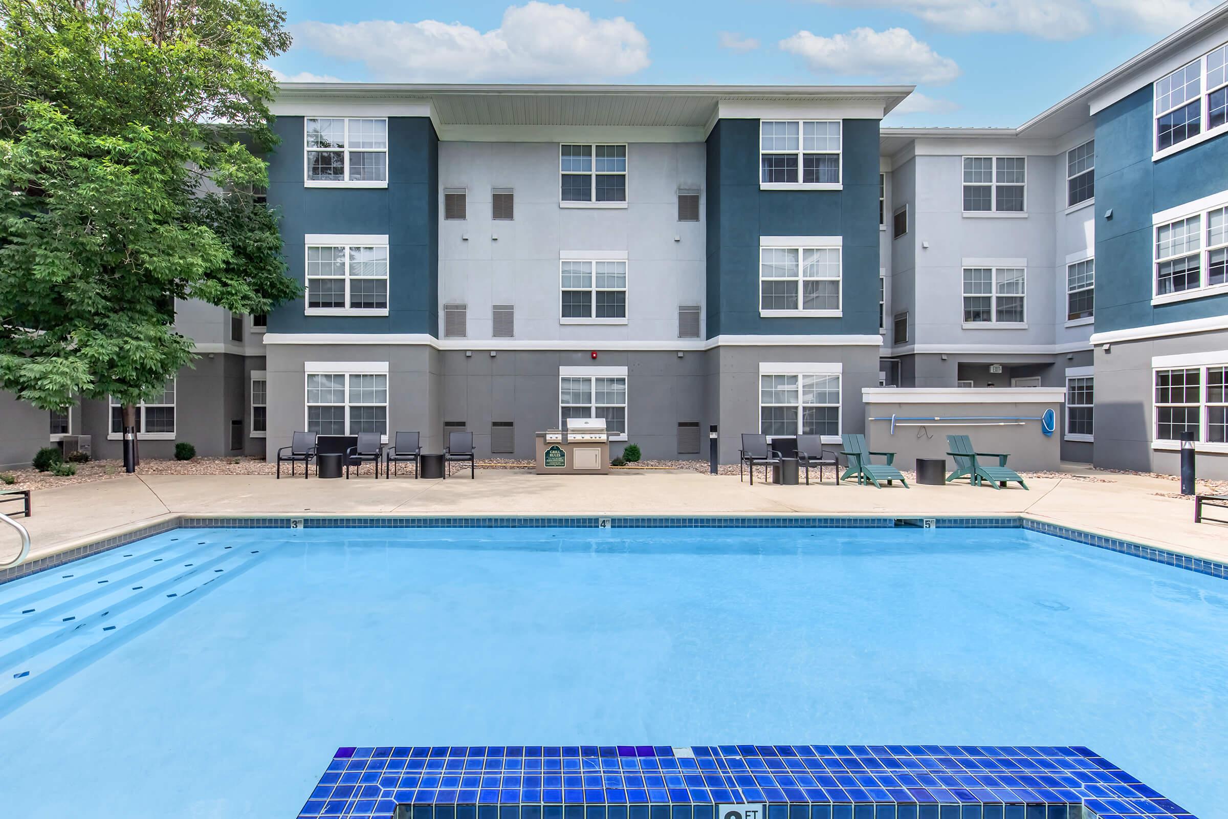 View of a swimming pool in front of a multi-story apartment building. The pool is surrounded by a patio area with lounge chairs and tables. The building features a modern design with blue and gray colors, and there are trees nearby providing shade. The sky is clear with a few clouds.