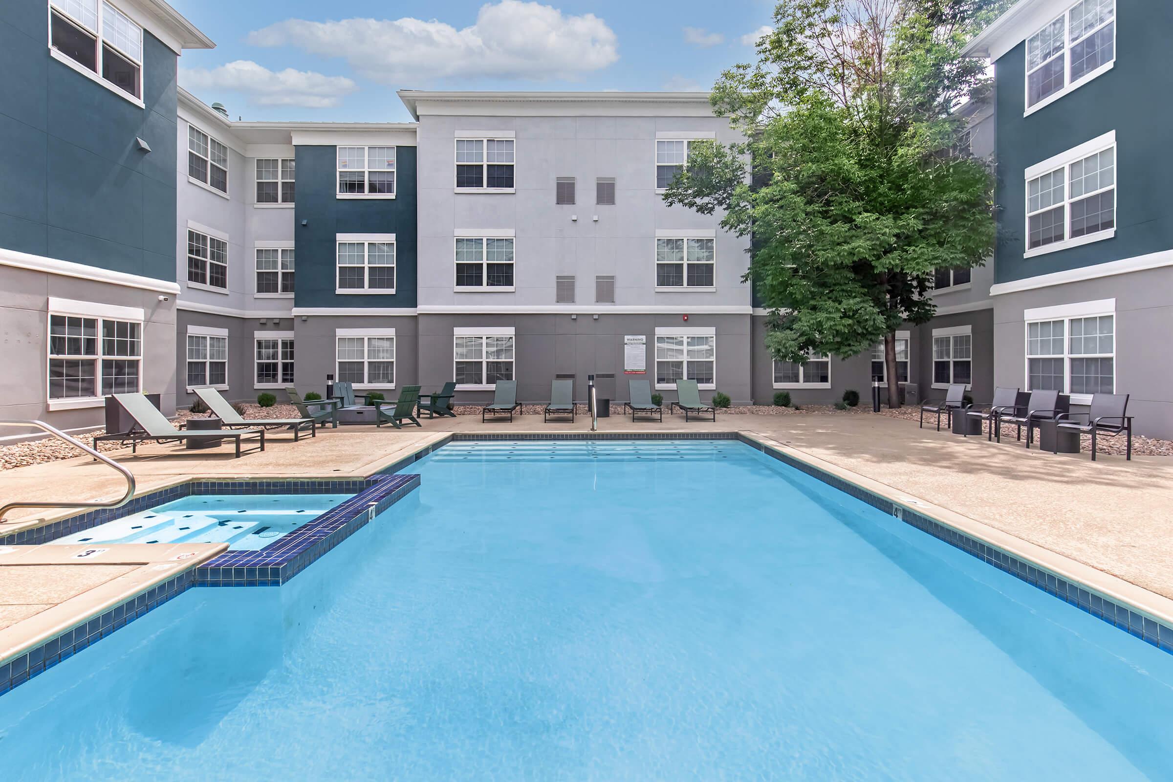 A clear blue swimming pool surrounded by lounge chairs, with a backdrop of a multi-story building featuring large windows. Lush trees provide greenery, and a bright sky can be seen above, creating a serene outdoor environment for relaxation.