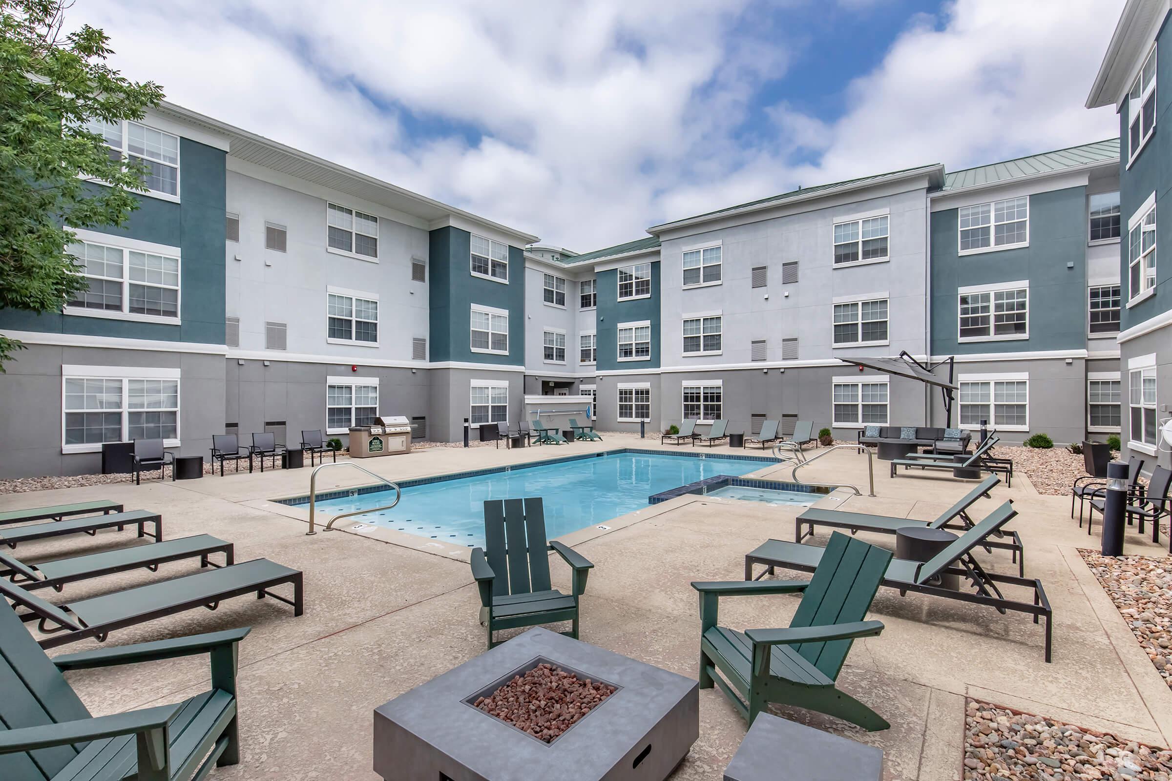 A modern apartment courtyard featuring a swimming pool surrounded by lounge chairs. There are fire pits and outdoor seating areas, framed by well-maintained greenery and multi-story buildings in the background under a partly cloudy sky.