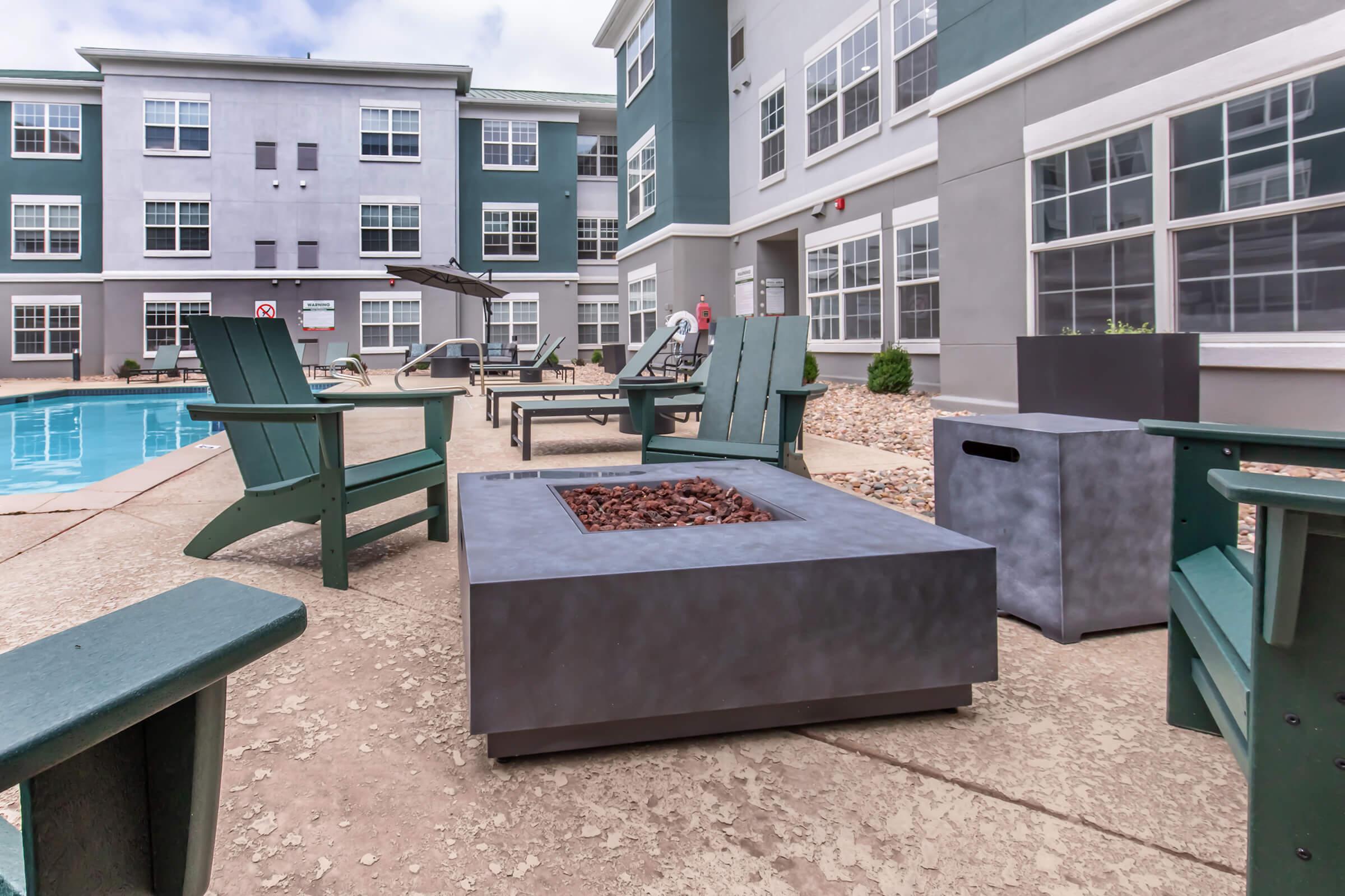 A modern outdoor lounge area featuring green Adirondack chairs arranged around a fire pit. The setting includes a swimming pool in the background, surrounded by stylish furniture and a well-maintained building with large windows. The scene is bright and inviting, perfect for relaxation.
