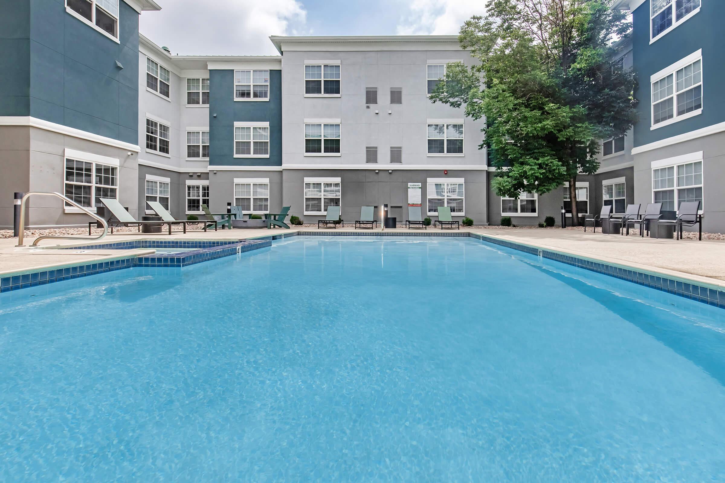 A tranquil swimming pool with clear blue water, surrounded by a patio area with lounge chairs and tables. The background features a modern multi-story building with large windows. Lush trees provide some greenery, enhancing the inviting atmosphere of the pool area.