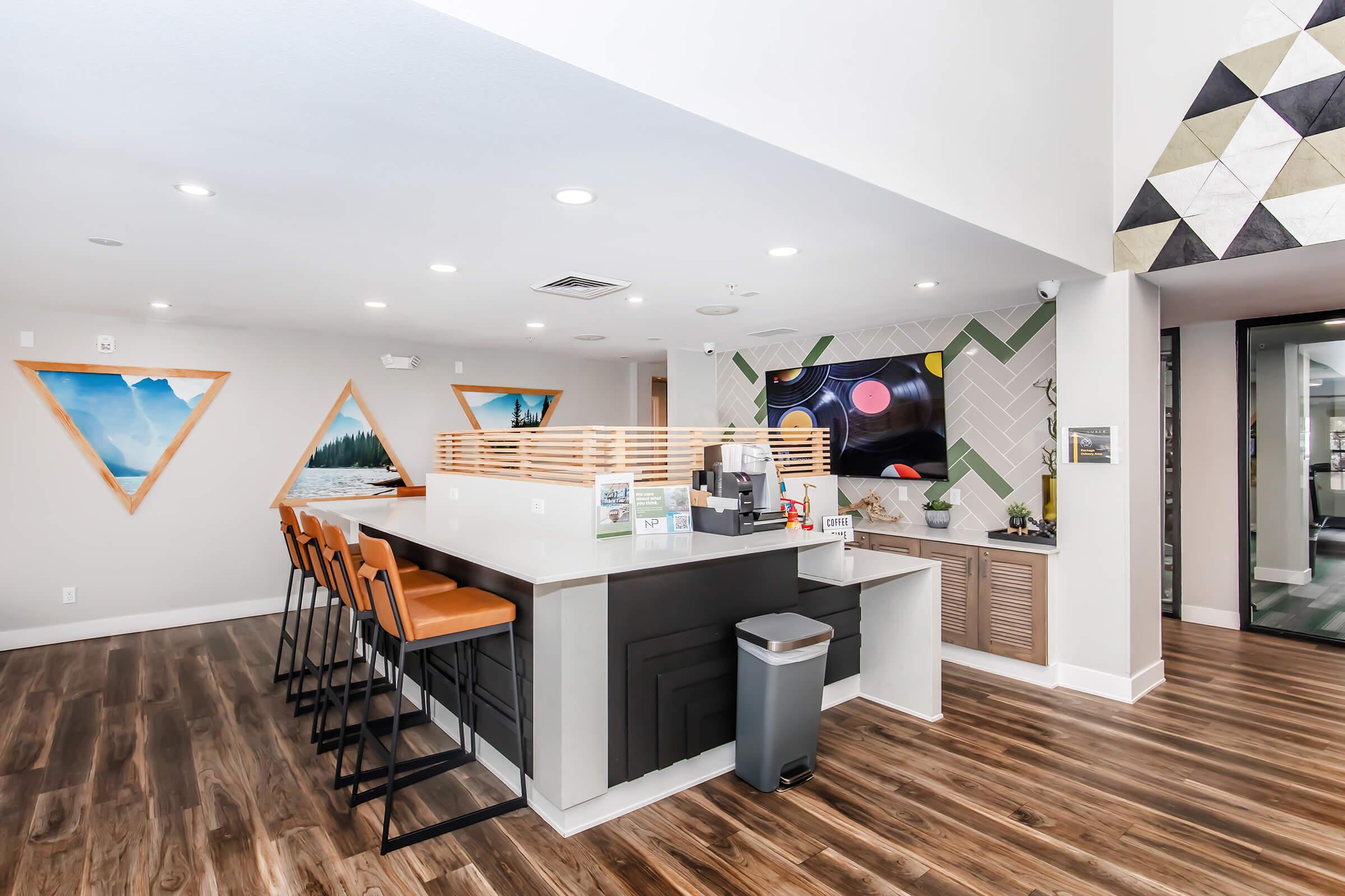 Modern reception area with a sleek front desk, high chairs, and a geometric accent wall. Decorative triangular wall art in shades of blue and orange adds a pop of color. The space features warm wooden flooring and a contemporary design, creating an inviting atmosphere for visitors.