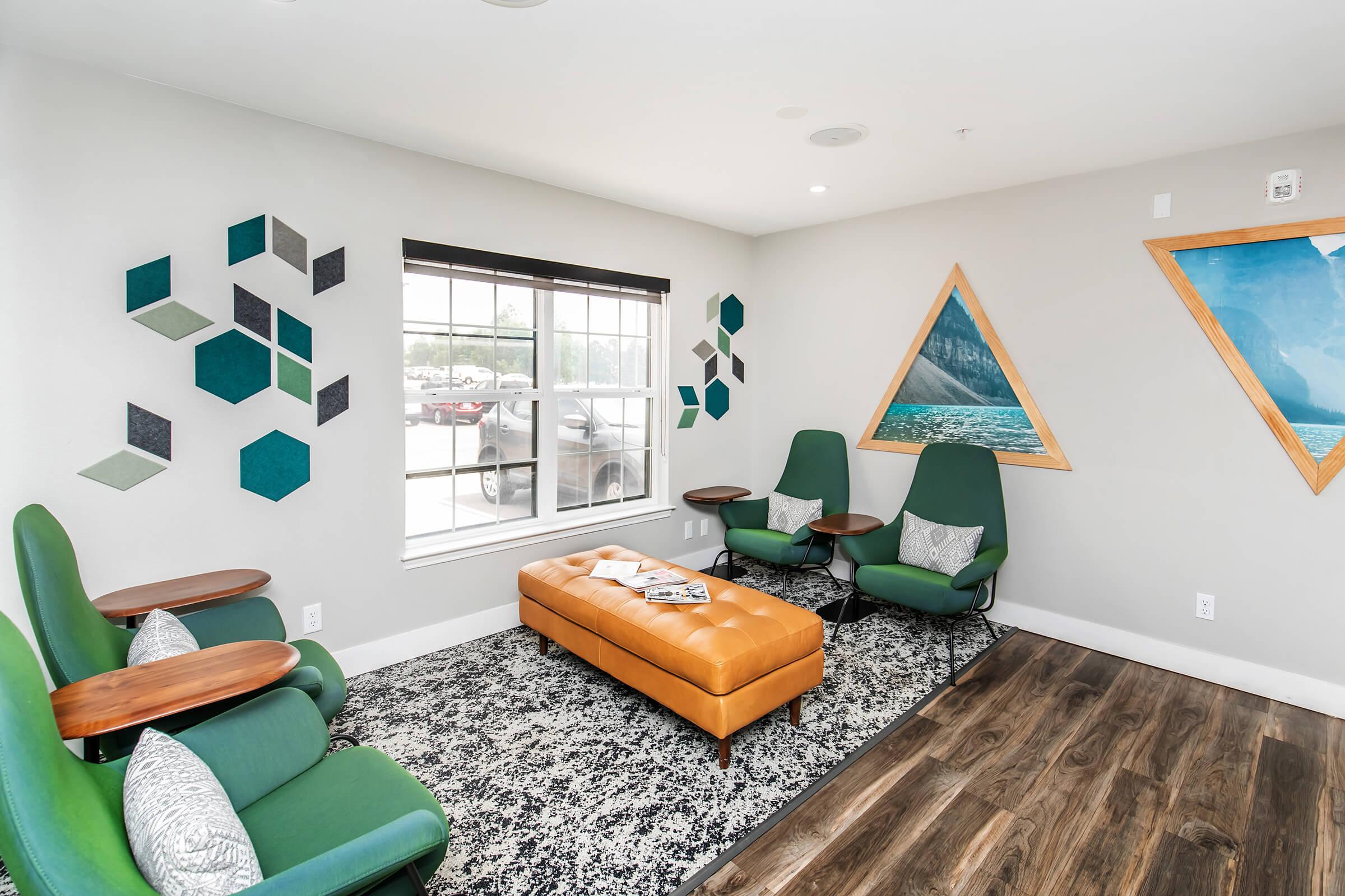 A modern lounge area featuring green chairs with wooden side tables, an orange couch, a patterned rug, and decorative wall art with hexagonal shapes and mountain landscapes. Natural light streams through a window, creating a welcoming atmosphere.