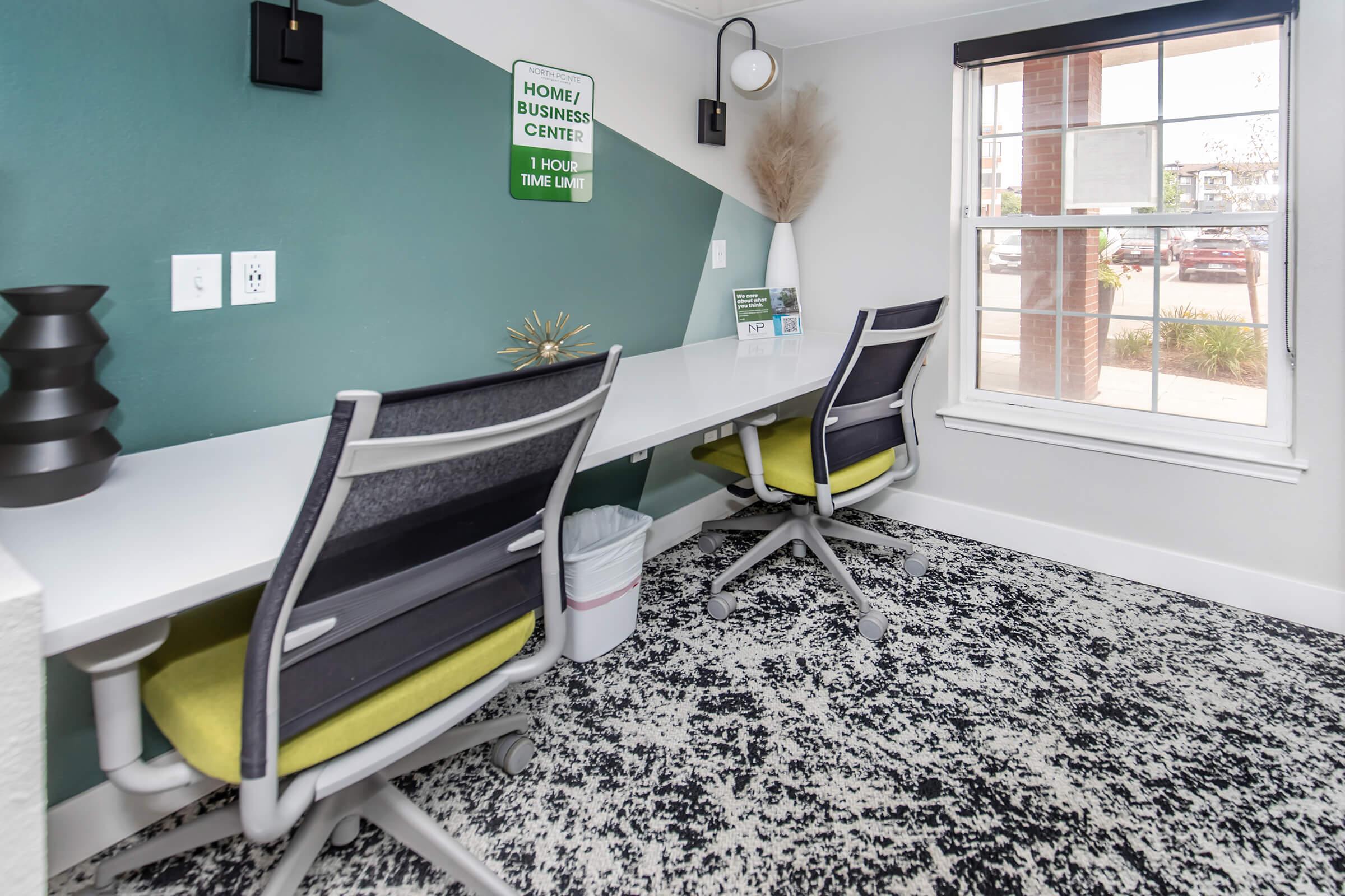 A modern workspace featuring two ergonomic chairs with bright green accents at a long white desk. The wall has a green and white geometric design, with decorative elements including a vase and a sunburst sculpture. A window provides natural light, and a sign indicates the space is for home or business use.