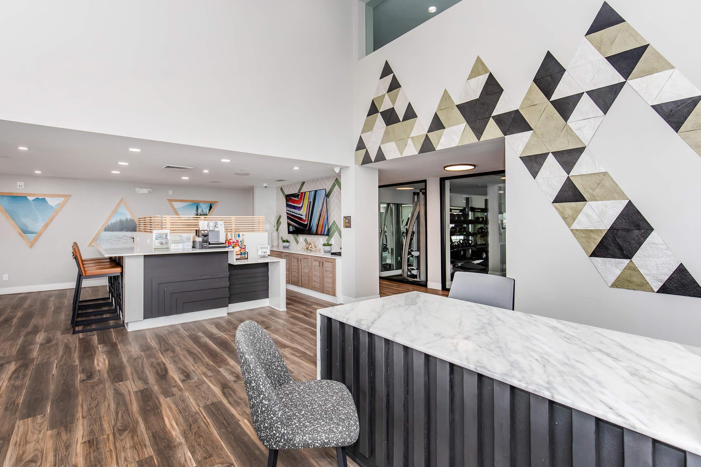 Modern interior of a reception area featuring a marble countertop, geometric wall designs in muted colors, and sleek wooden flooring. The space includes a front desk, seating area, and fitness room visible through glass doors, creating a contemporary and inviting atmosphere.