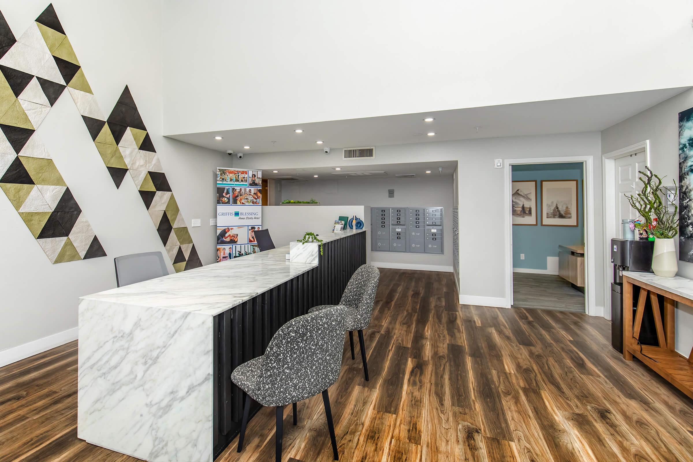 Modern reception area featuring a marble front desk, stylish gray chairs, and a decorative wall with geometric patterns. There are also lockers for storage and a welcoming ambiance with wooden flooring and neutral-colored walls.