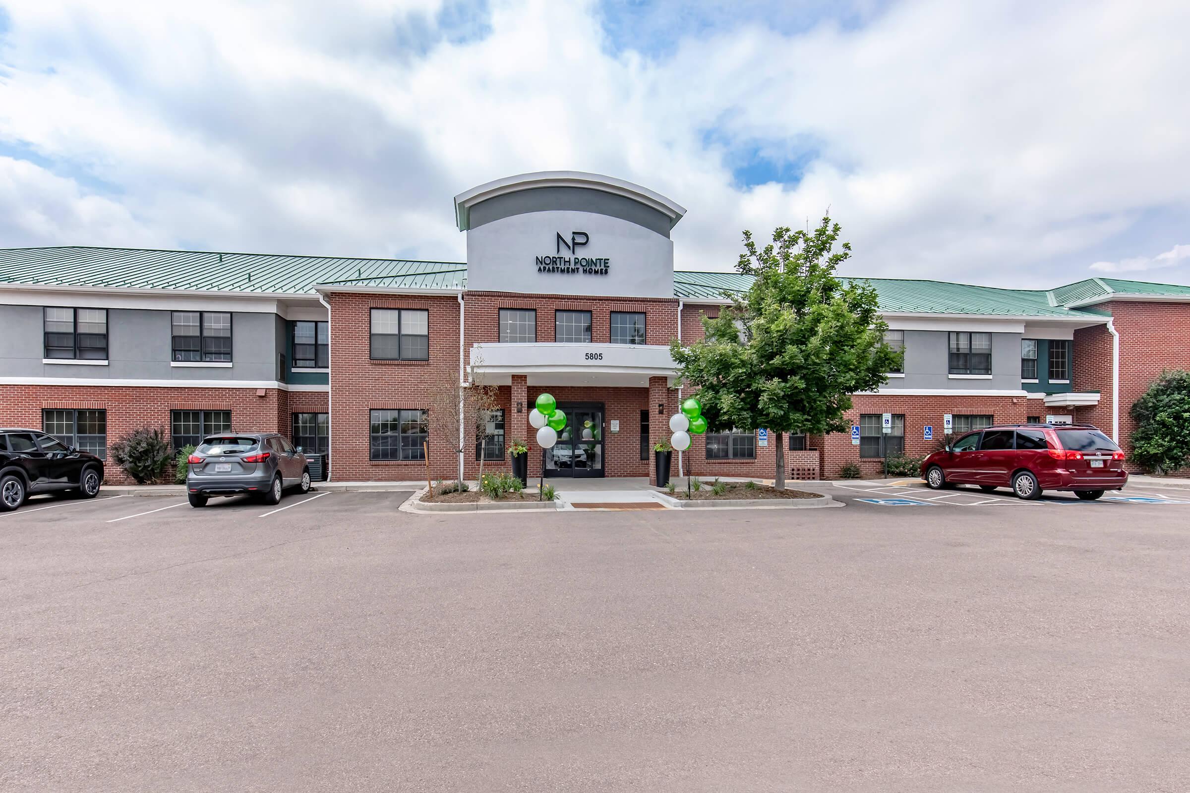 Exterior view of North Pointe Health Center, a two-story brick building with a green roof. The entrance features a covered awning with balloons in green and white, and parking spaces are visible in front. The surrounding area includes landscaped trees and shrubs.