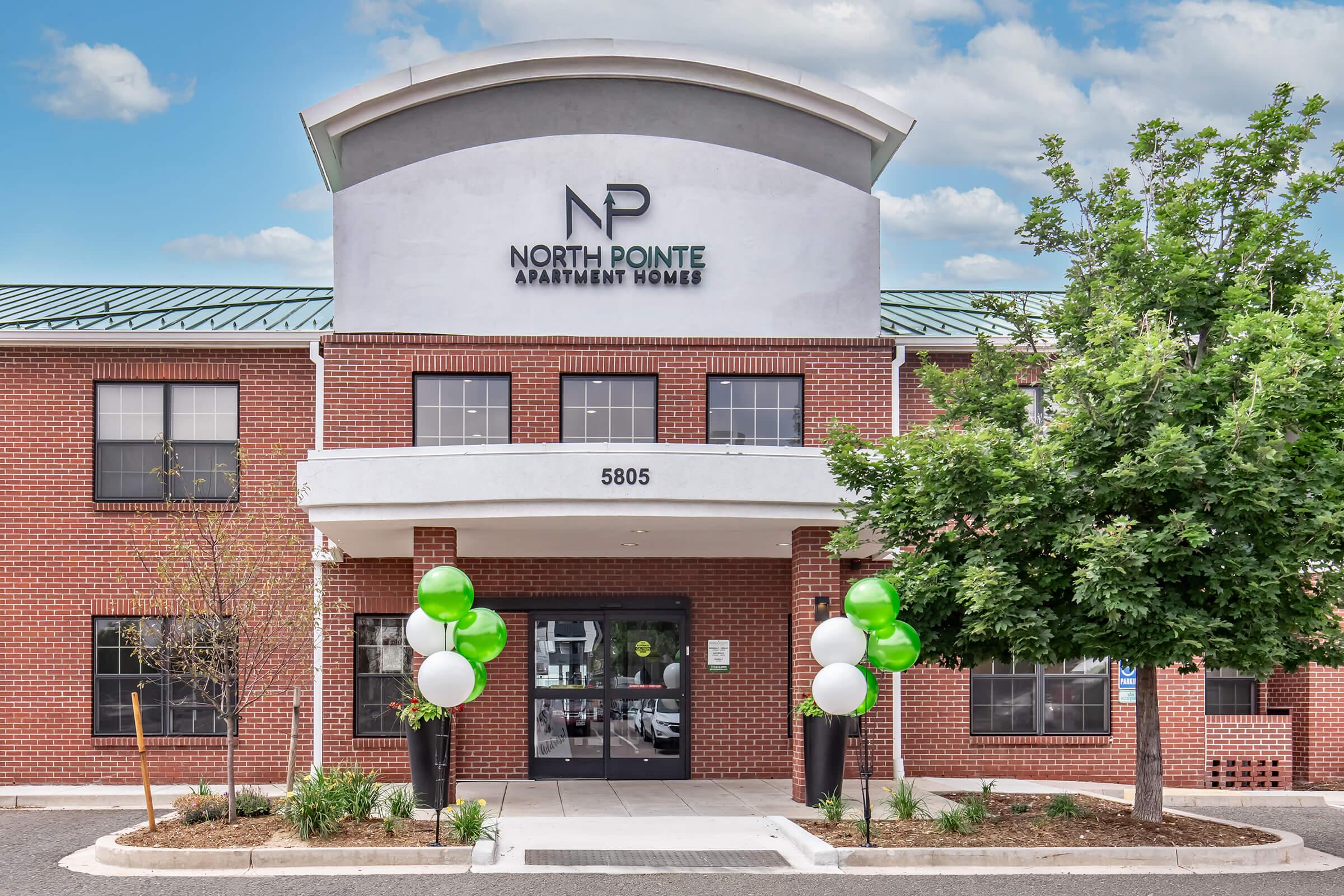 Exterior view of North Pointe Apartment Homes, a two-story brick building featuring a prominent entrance with a curved awning, large windows, and a clean, modern design. Green and white balloons are displayed at the entrance, complementing the welcoming atmosphere.