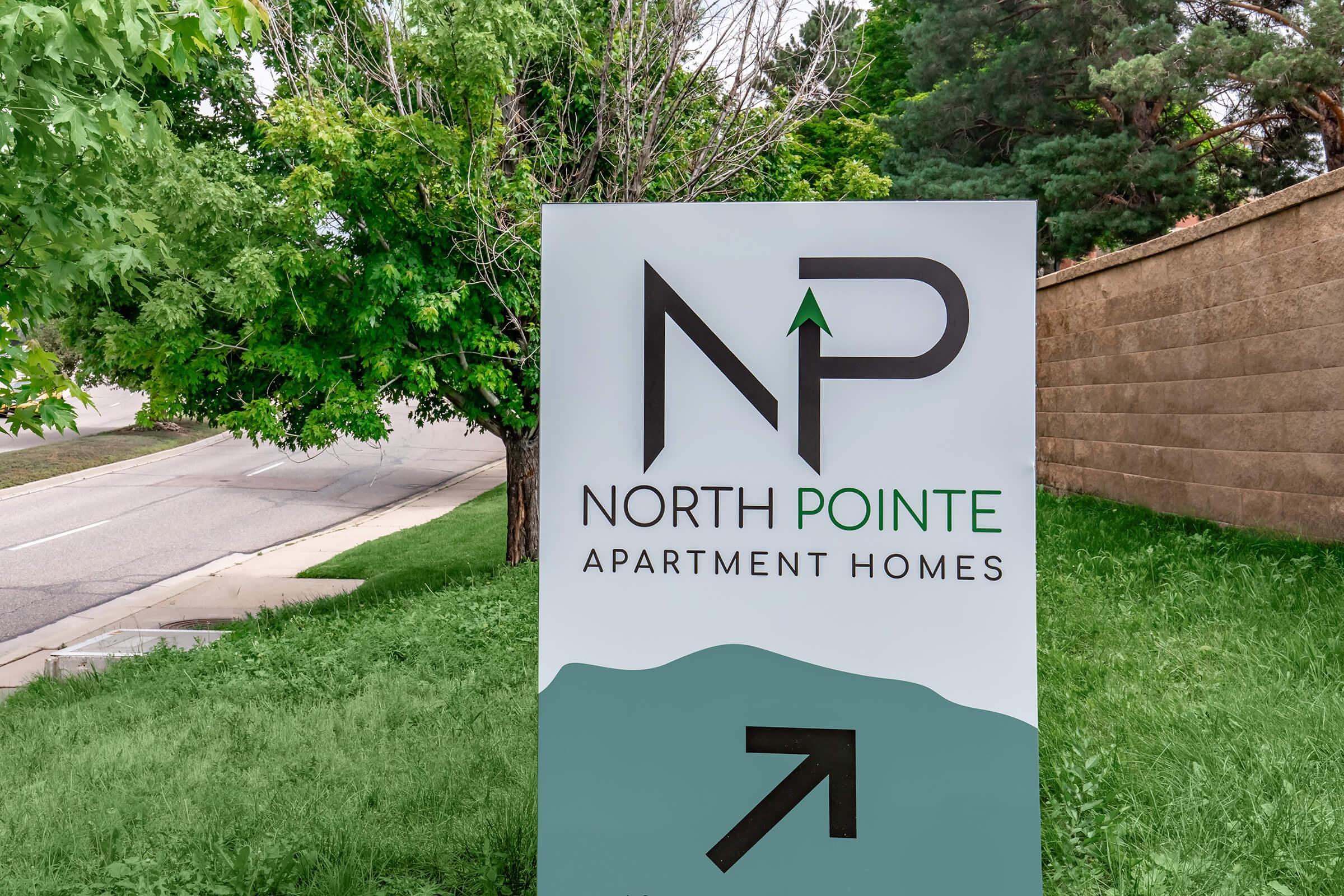 Sign for North Pointe Apartment Homes featuring the logo "NP" with a green mountain graphic and an arrow pointing right. The background includes greenery and a view of a road.