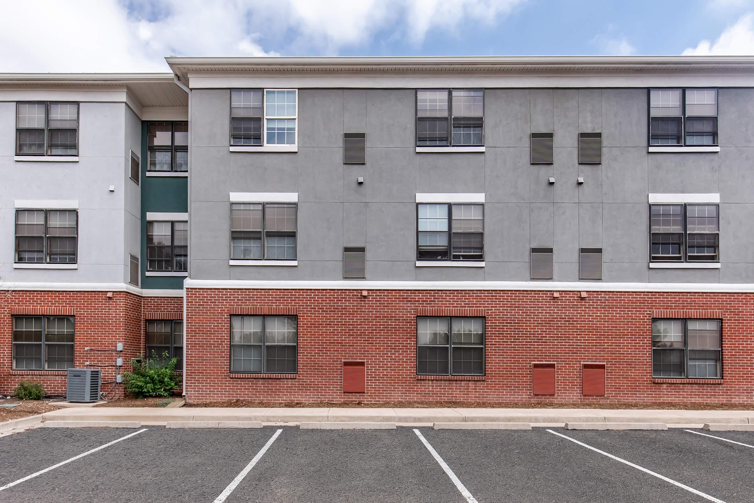 A three-story apartment building with a mix of gray and red brick exterior. The facade features multiple windows with black grilles and ventilation grates. A parking lot is visible in front, with marked spaces. Green landscaping is present at the base of the building. The sky is partly cloudy.