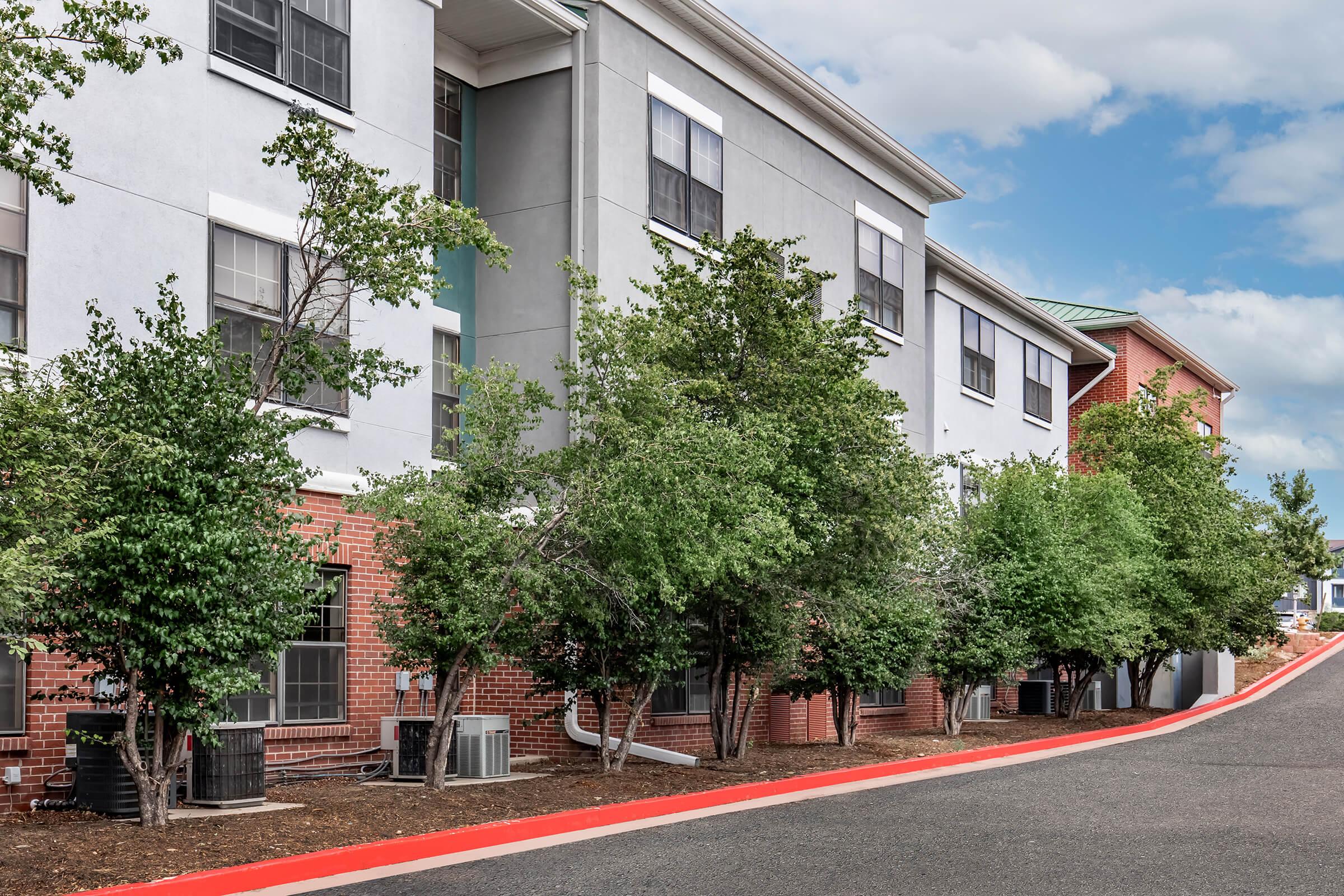 A modern multi-story building with a mix of gray and brick exteriors, surrounded by small trees and air conditioning units. The foreground features a red paved walkway, leading up to the building, under a partly cloudy sky.
