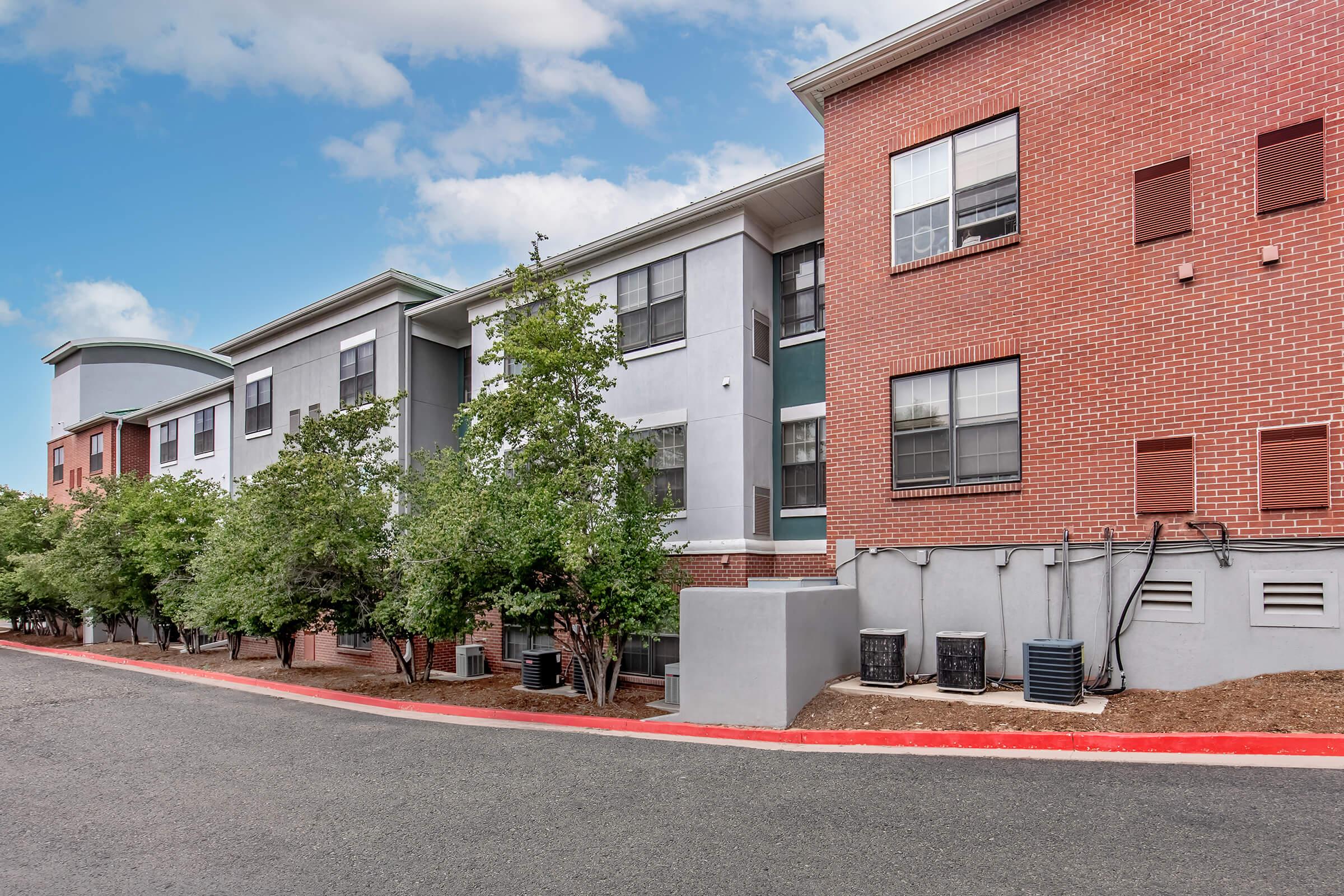A residential building with a mix of brick and gray exterior features. The structure includes multiple windows and air conditioning units. Lush green trees line the walkway, and there is a paved road with a red curb in the foreground, set against a partly cloudy blue sky.