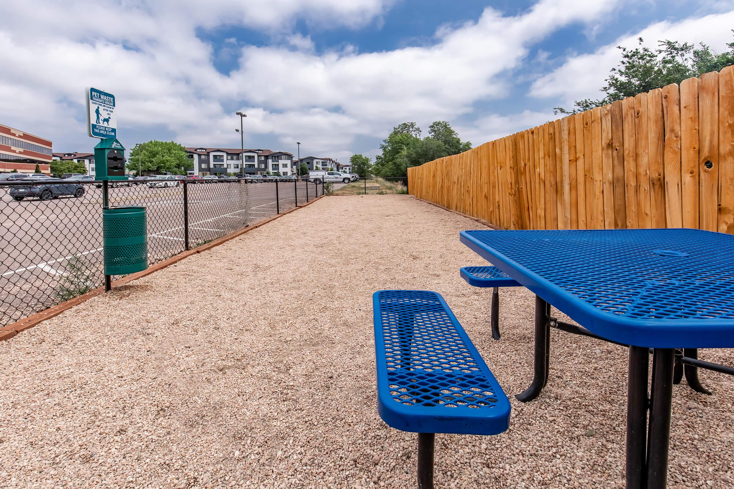 Blue picnic table on gravel surface near a wooden fence, with a grassy area and a sign in the background. The sky is partly cloudy, and there are a few buildings visible in the distance. The setting appears to be a park or recreational area.
